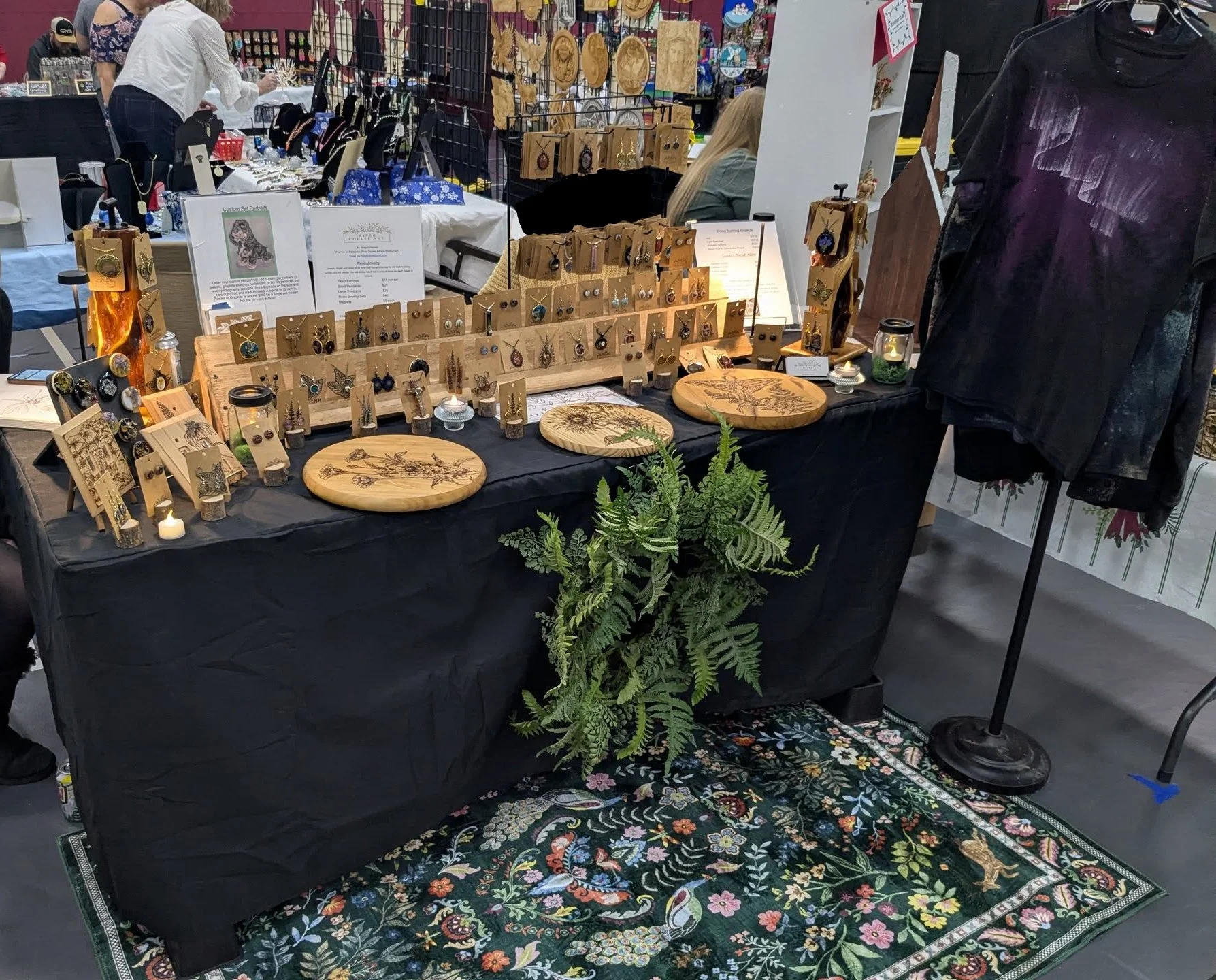 A vendor's booth displaying handcrafted jewelry and wood art, including earrings, necklaces, and paintings, at an indoor market. The booth has a black tablecloth, a patterned rug underneath, and various plants as decoration.