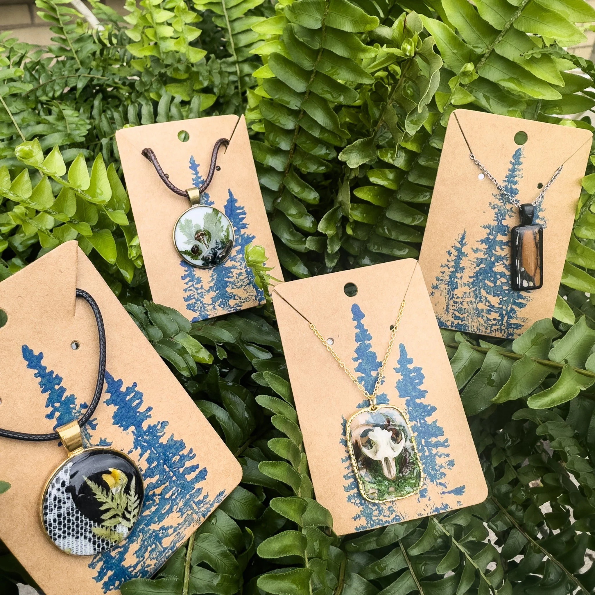Four jewelry necklaces displayed on cardboard cards with blue tree designs, set outdoors on green foliage.
