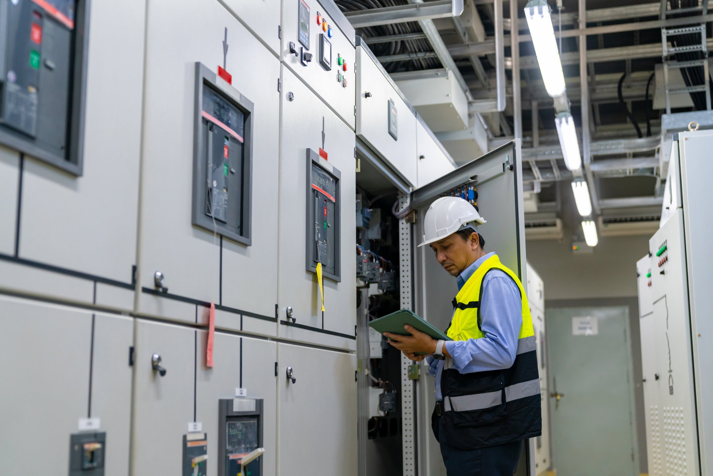 A man in a hard hat and yellow safety vest inspecting electrical panels in an industrial facility.