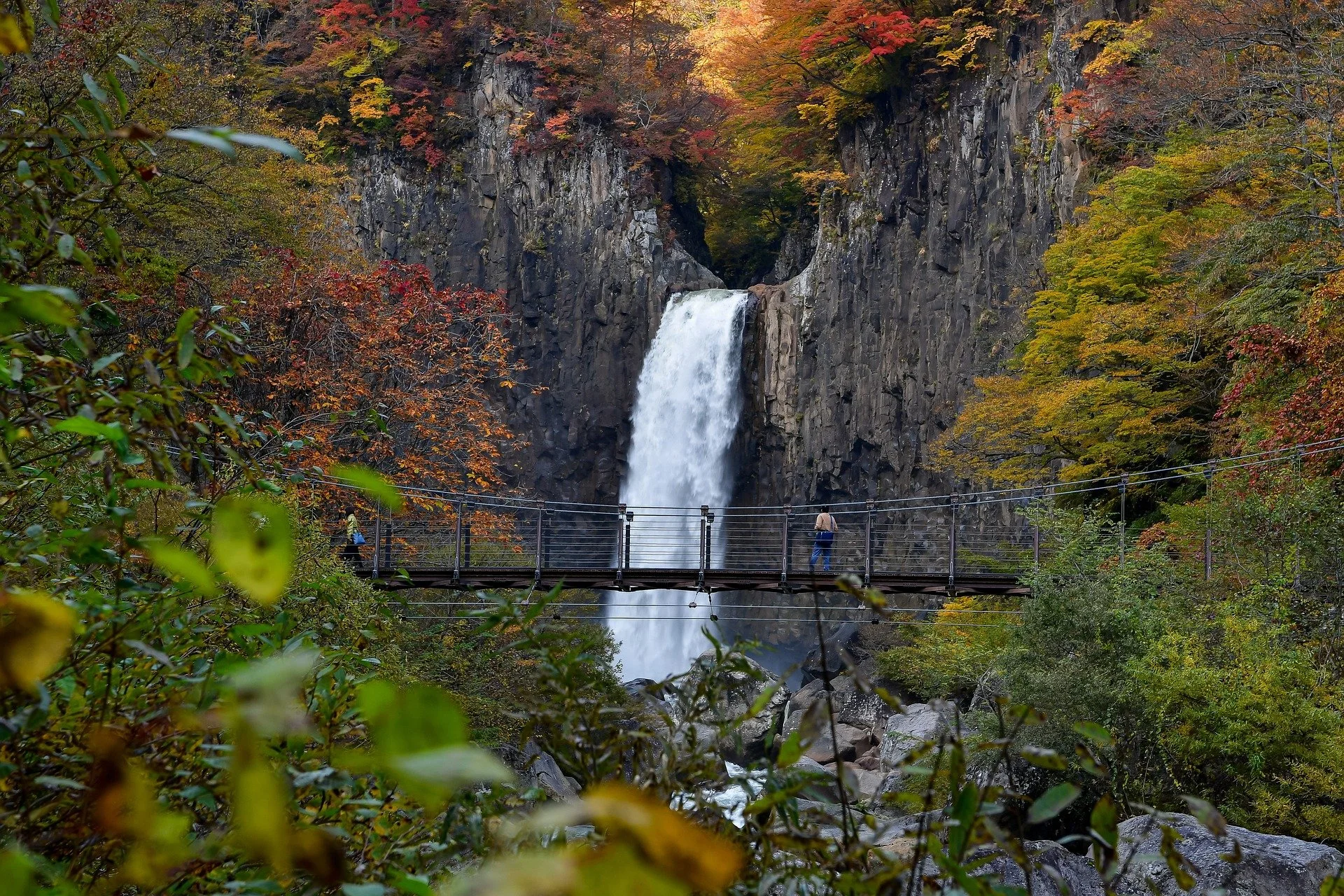 A waterfall in a forest with fall foliage, viewed from a bridge with people walking across.