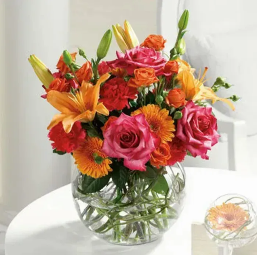 Colorful bouquet of pink, orange, and yellow flowers in a clear glass vase on a white table.