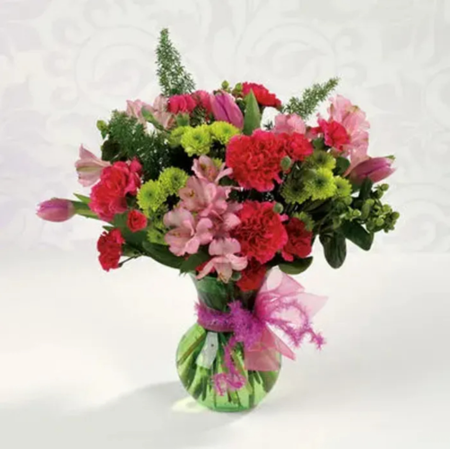 A colorful bouquet of pink, red, and green flowers in a striped green vase with pink decorative wrapping.
