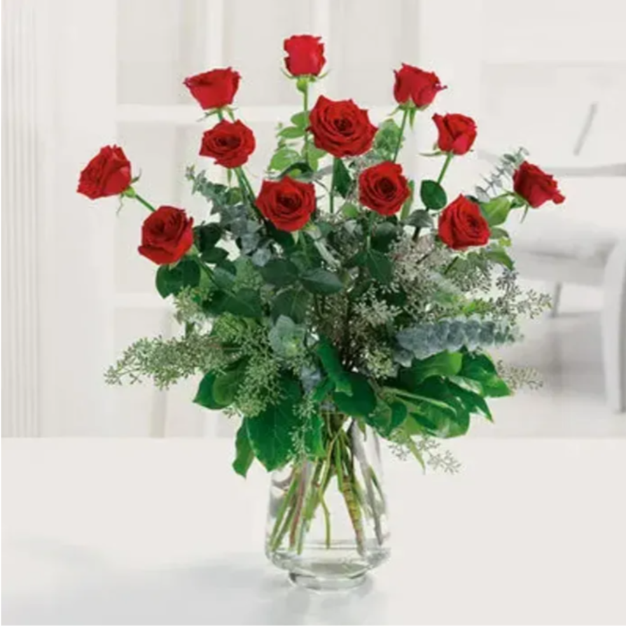 Vase of red roses and assorted greenery on a white surface.