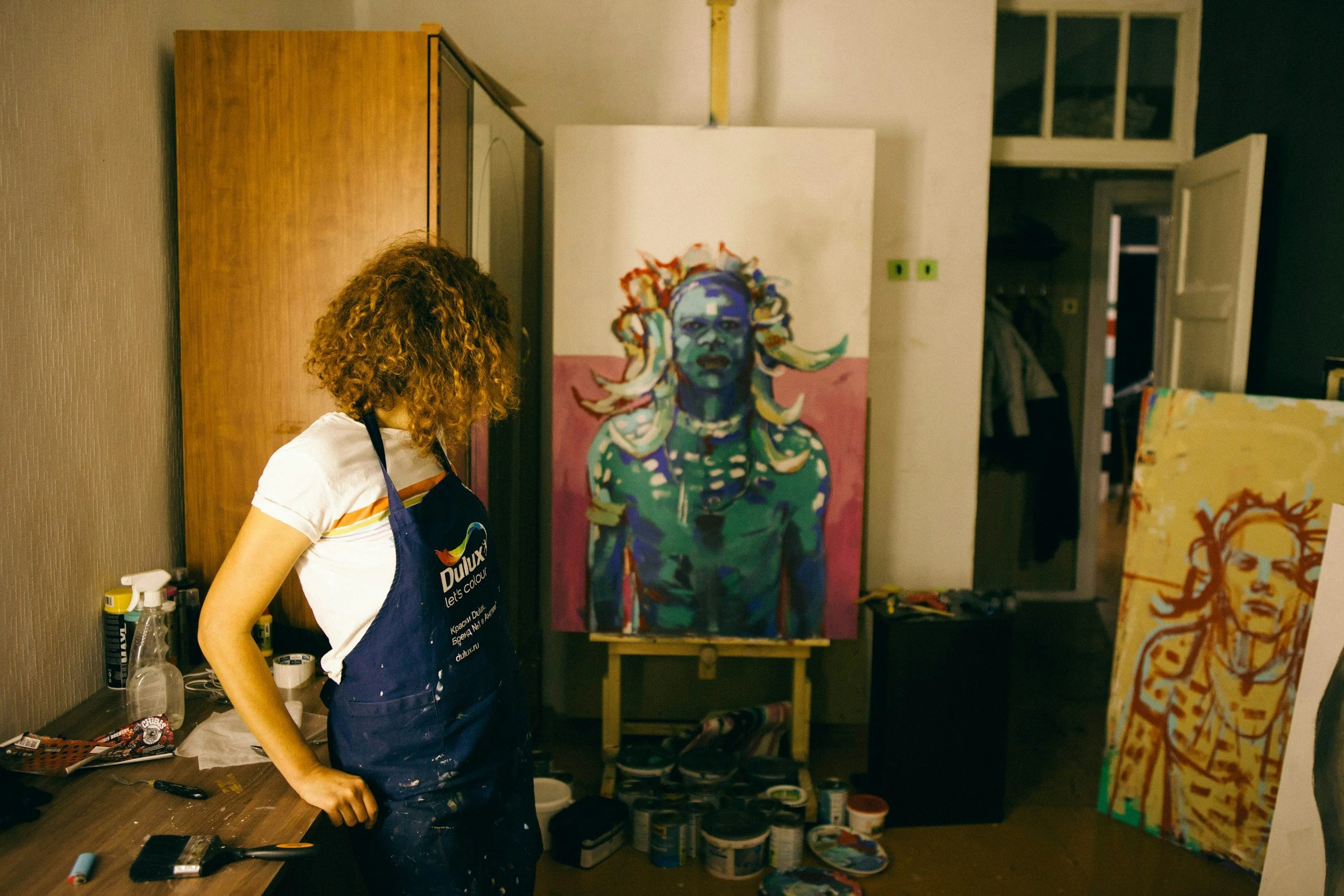 An artist with curly hair wearing a Dulux apron stands in her studio, observing a colorful portrait of a woman with wild hair and intense expression. The studio contains various art supplies, paintings, and an open door leading to a closet.