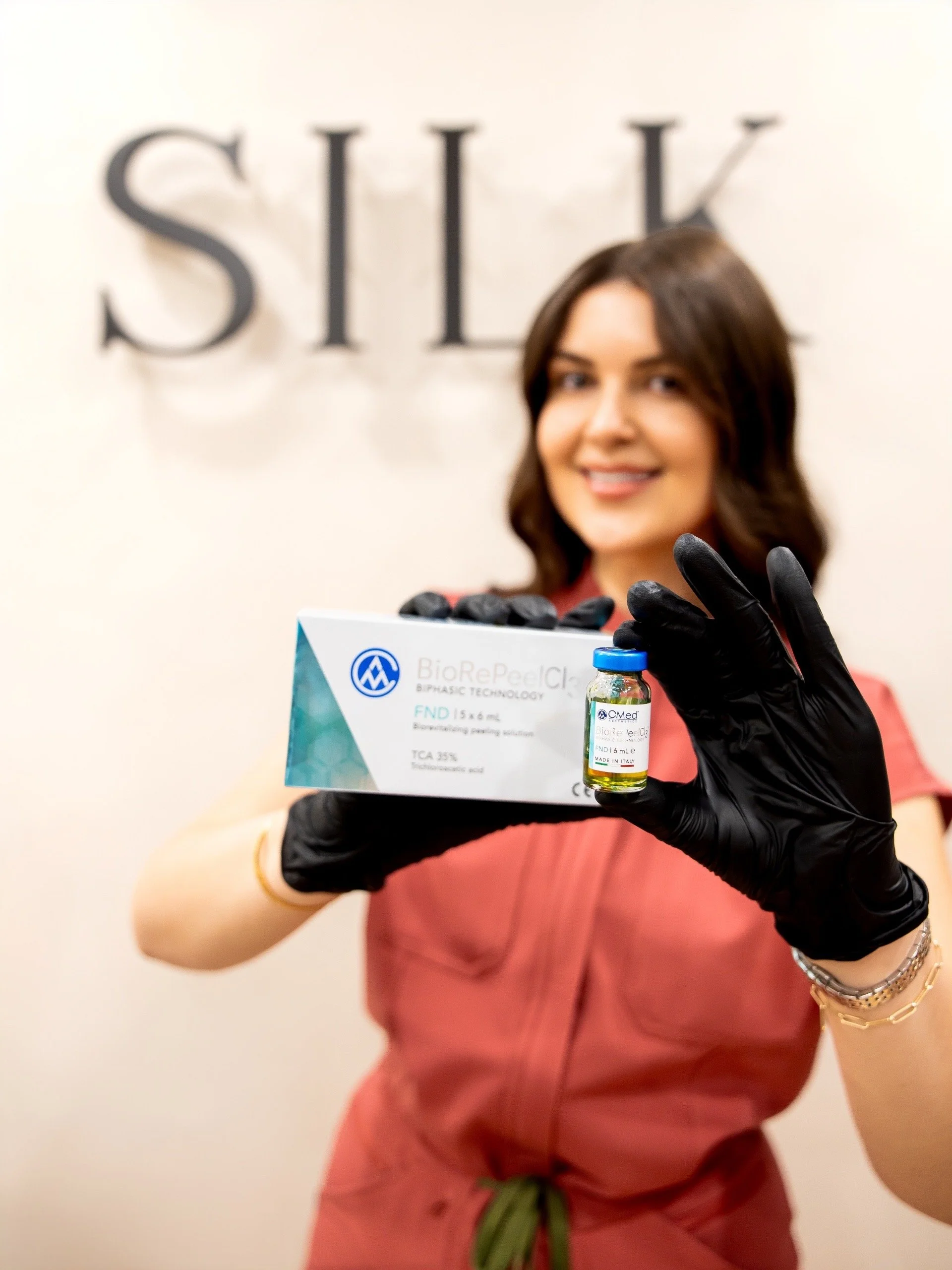 A smiling aesthetician in black gloves holds a BioRePeelCl3 chemical peel box and vial.