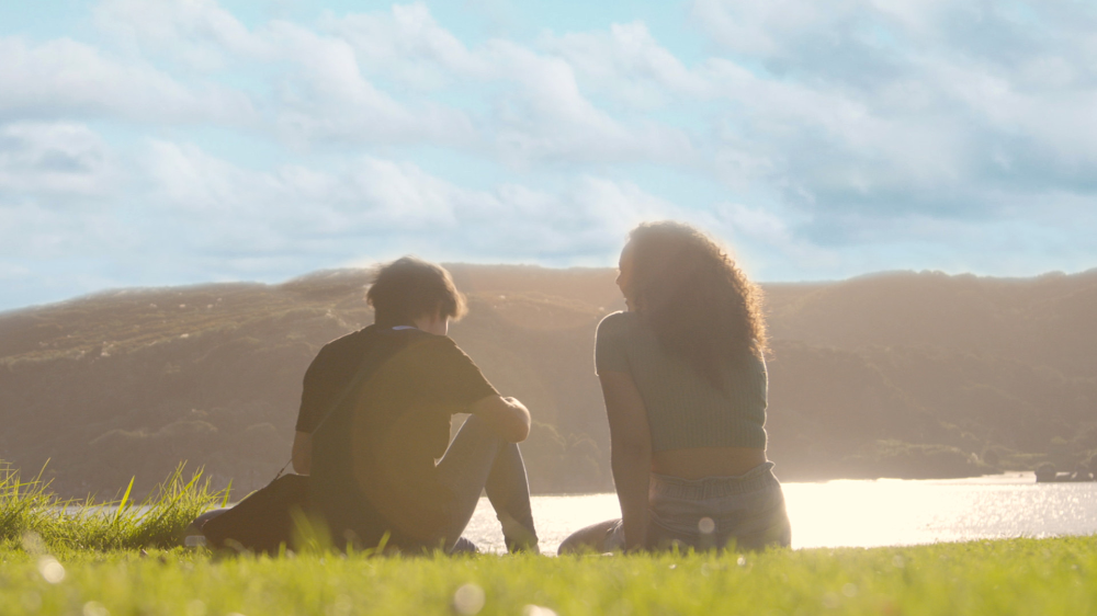 Two people sitting on grass near a body of water under a blue sky with clouds, with hills in the background.