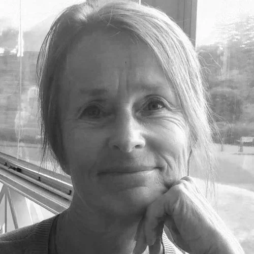 A woman with light-colored hair, smiling gently, resting her chin on her hand, sitting near a window with an outdoor view.