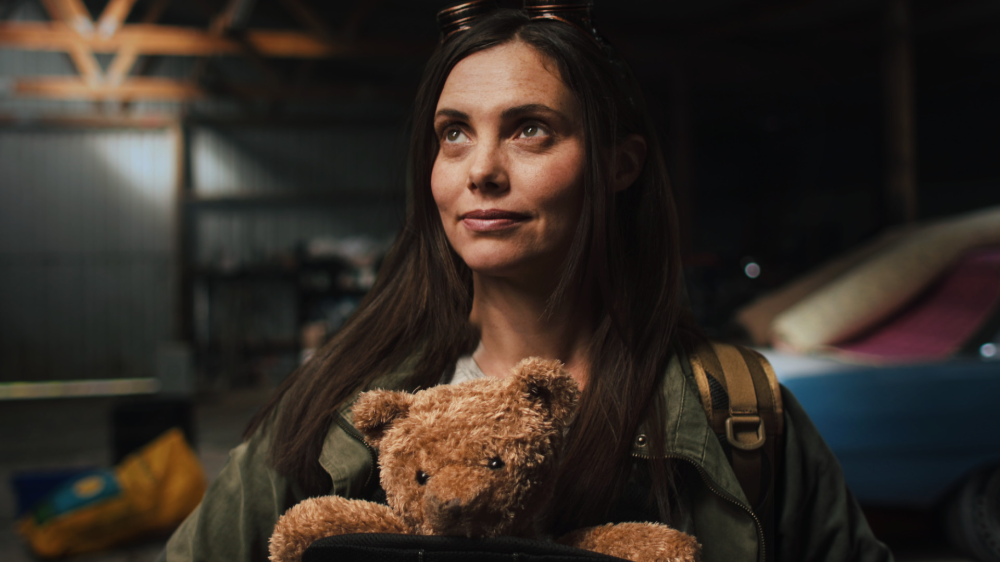 Woman with long brown hair and a backpack, holding a teddy bear, standing in a dimly lit warehouse or garage.