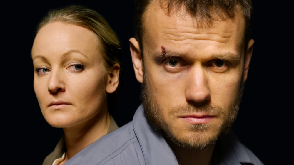A woman and a man with serious expressions look at the camera against a black background. The woman has blonde hair, and the man has a wound on his forehead.