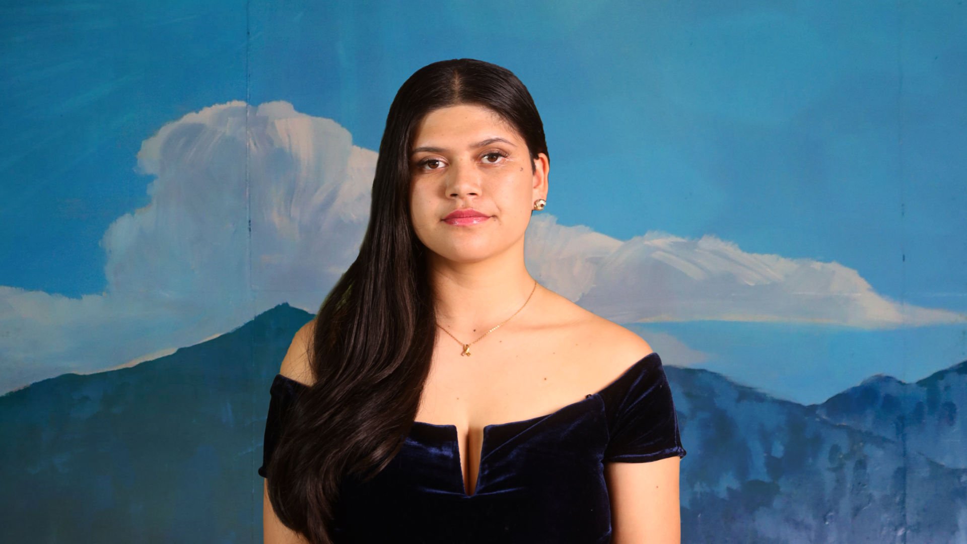 A woman with long dark hair wearing an off-the-shoulder navy blue dress and gold jewelry stands in front of a painted backdrop of blue mountains and a sky with clouds.