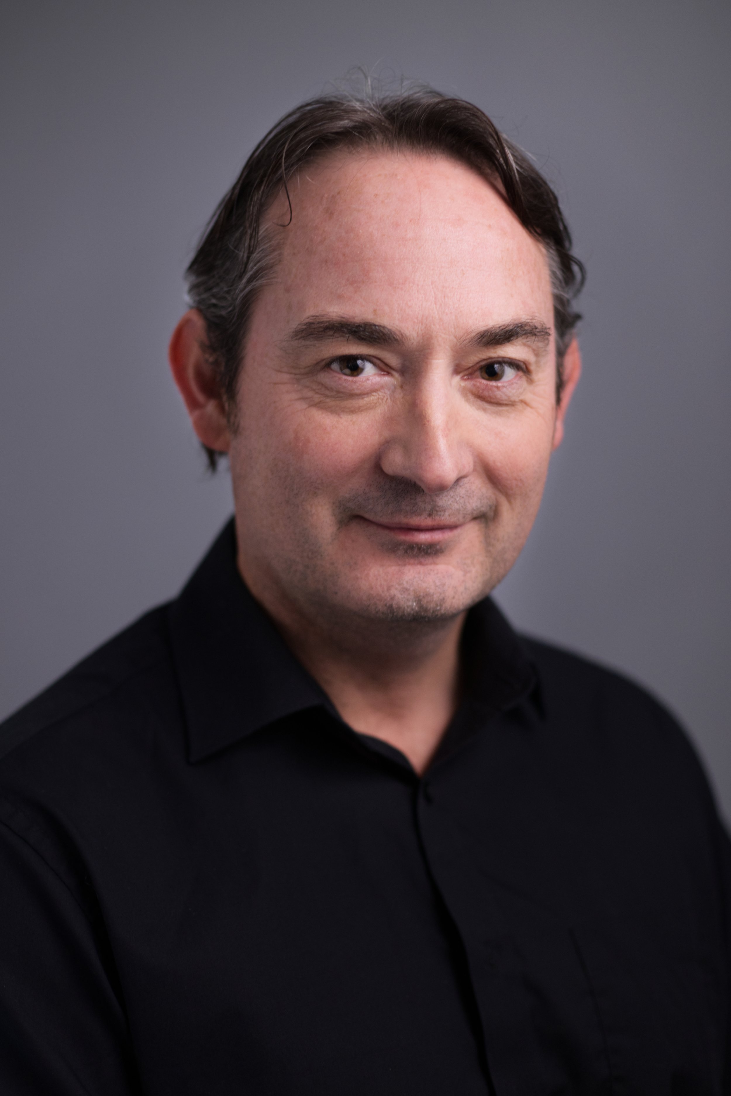 A man with dark hair, wearing a black shirt, smiling at the camera against a gray background.