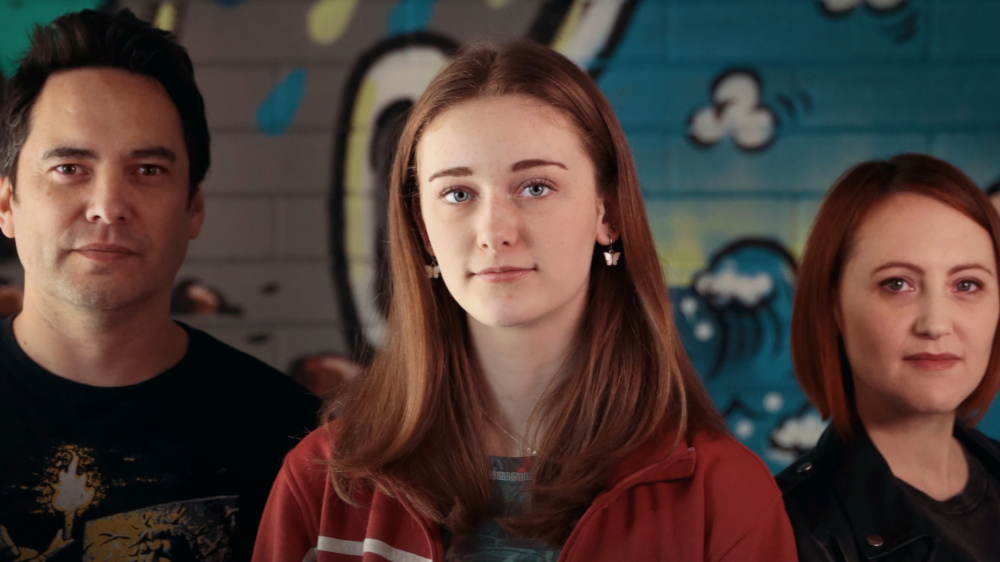 Three people, two women and one man, pose in front of a colorful graffiti wall. The woman in the center has long red hair and wears a red jacket. The man on the left has short dark hair and wears a black shirt. The woman on the right has shoulder-length red hair and wears a dark jacket.