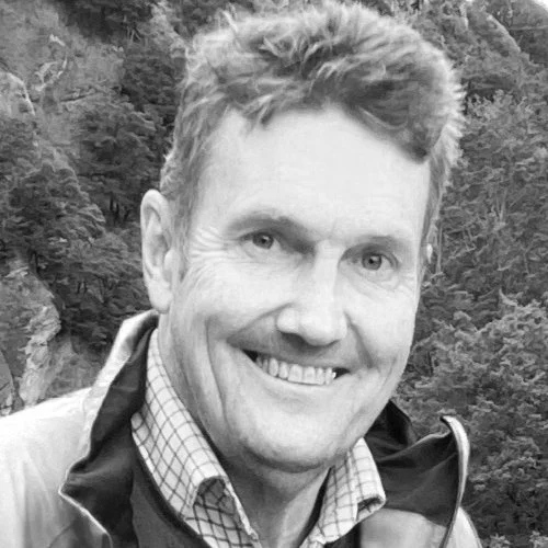 A man smiling outdoors with trees and rocks in the background.