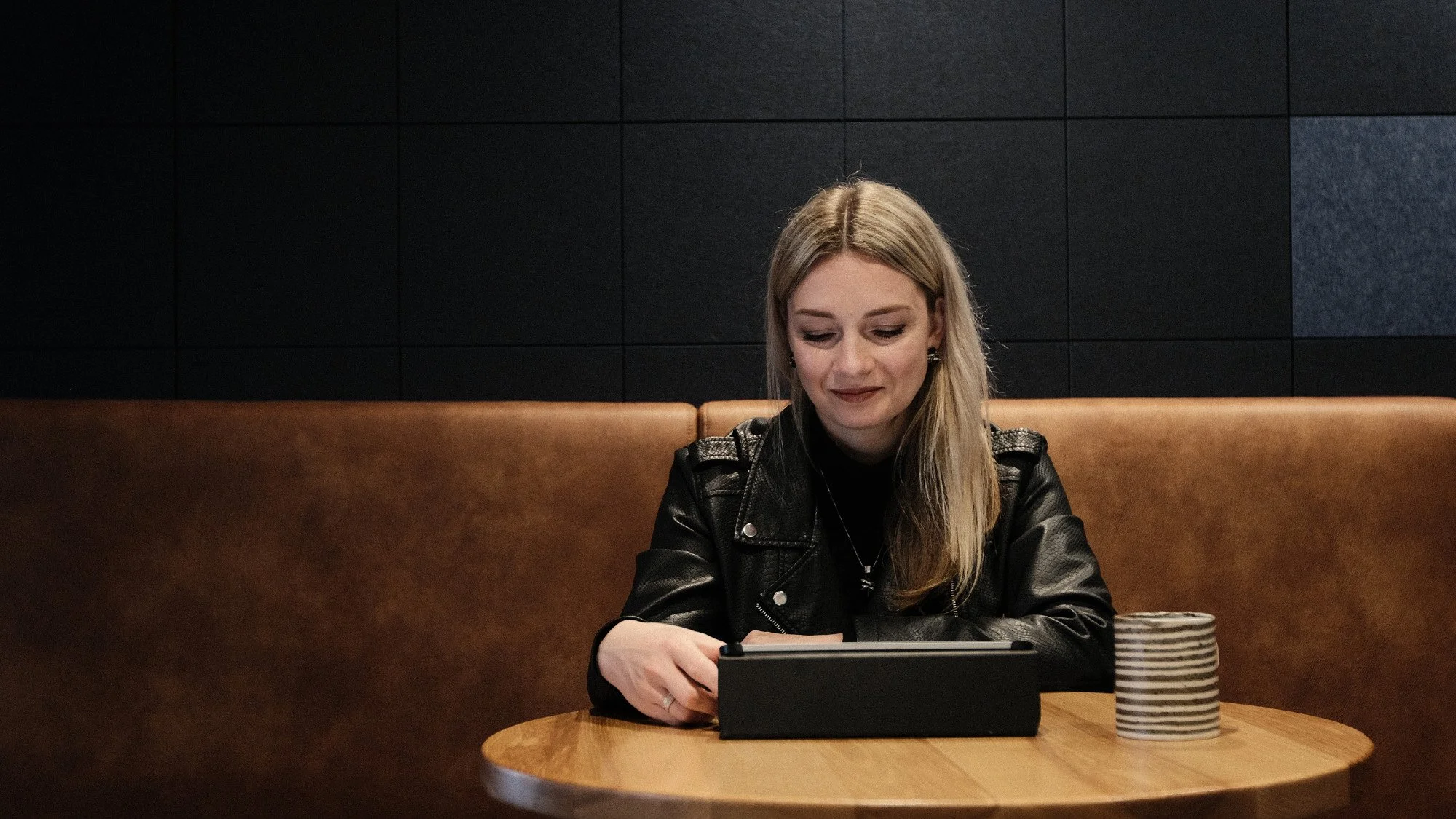 A woman with long blonde hair wearing a black leather jacket sitting at a wooden table, looking at and smiling at a tablet device. A striped ceramic cup is on the table to her right. The background features a dark tiled wall.