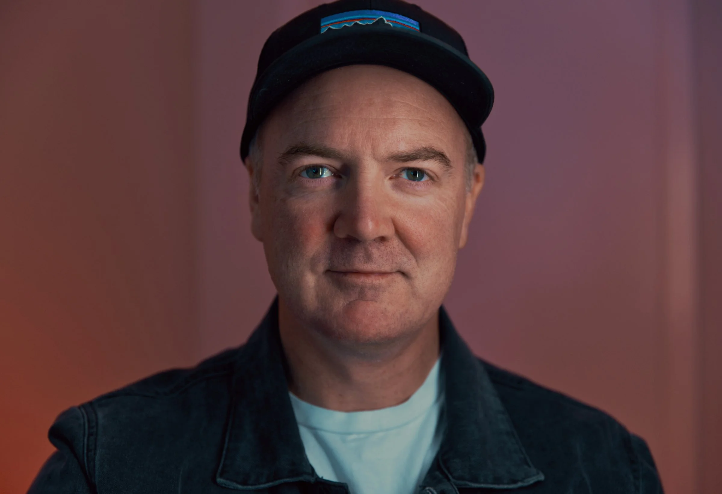 A middle-aged man with blue eyes, wearing a black cap with a colorful logo and a denim jacket, standing in front of a pinkish background.