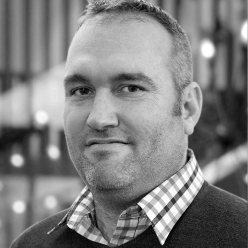 A grayscale portrait of a man with short hair, wearing a checkered shirt and a dark sweater, standing outdoors with blurred background lights.