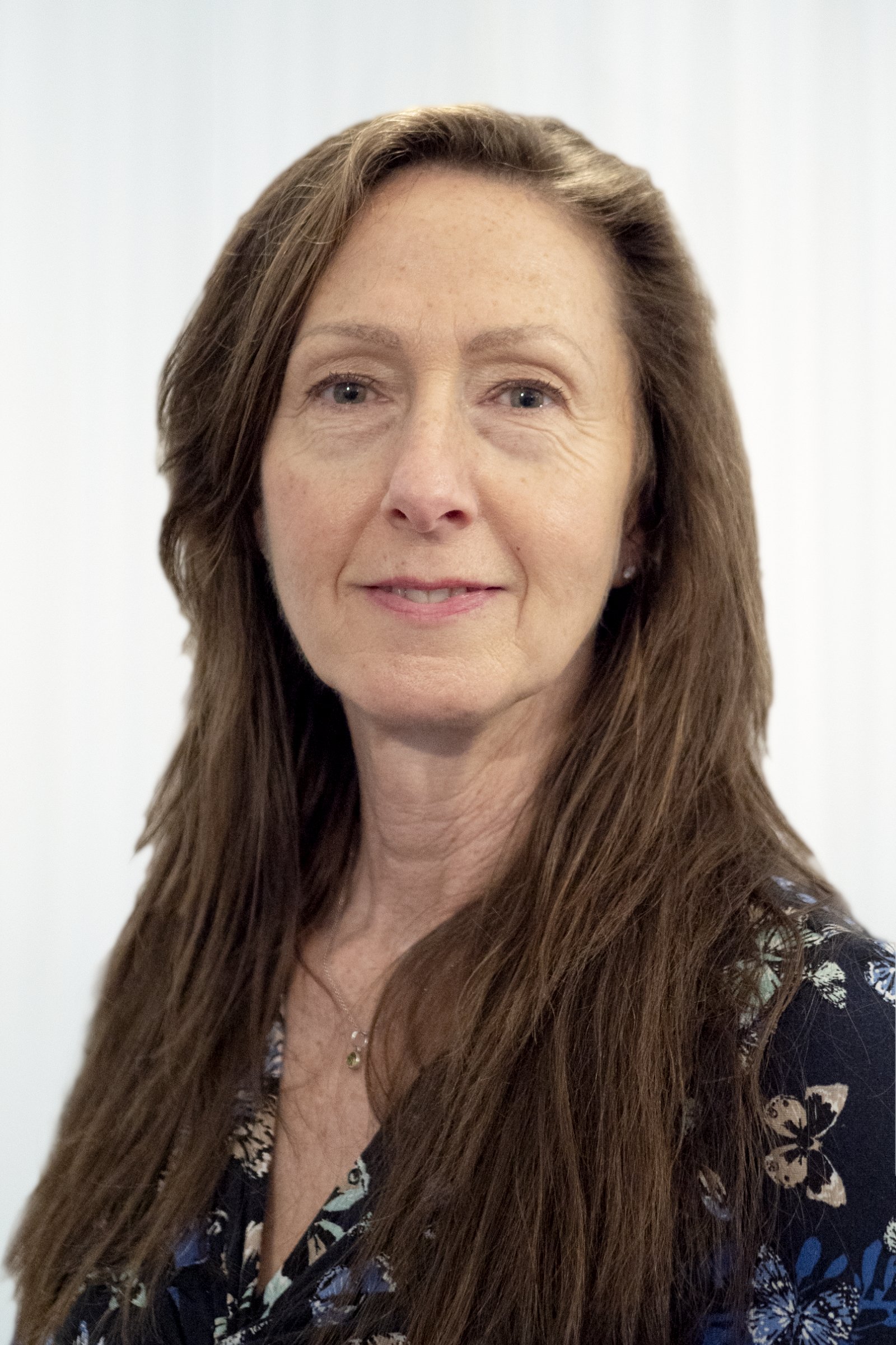 A portrait of a woman with long brown hair, wearing a dark floral top, standing against a plain white background.