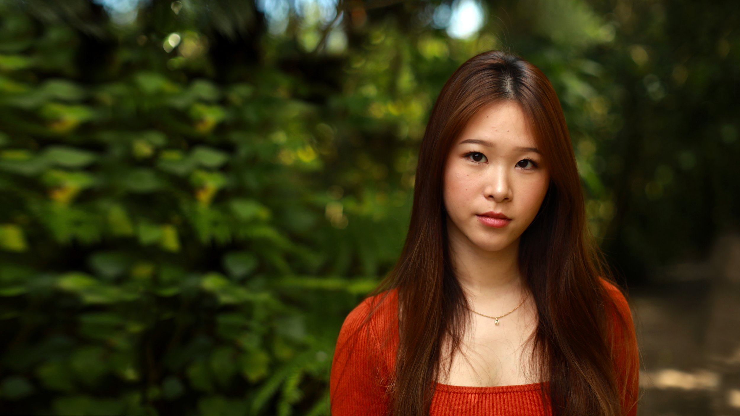 Young woman with long brown hair wearing an orange top standing outdoors with green foliage in the background.