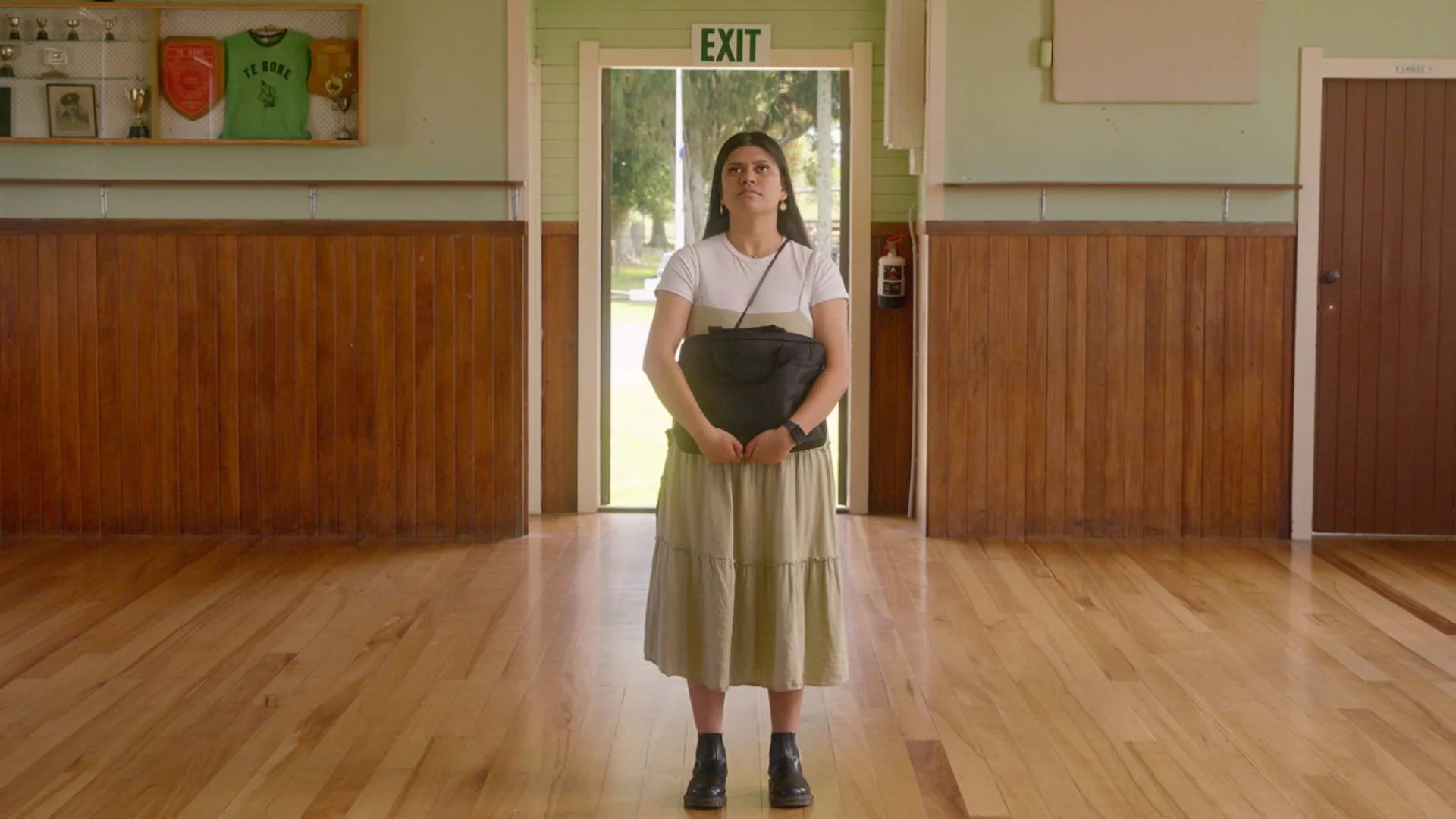 A woman standing in a room with wooden flooring, holding a black bag, looking upward. Behind her, a glass door with an 'EXIT' sign above. The room has wood-paneled walls with sports memorabilia displayed on a shelf.