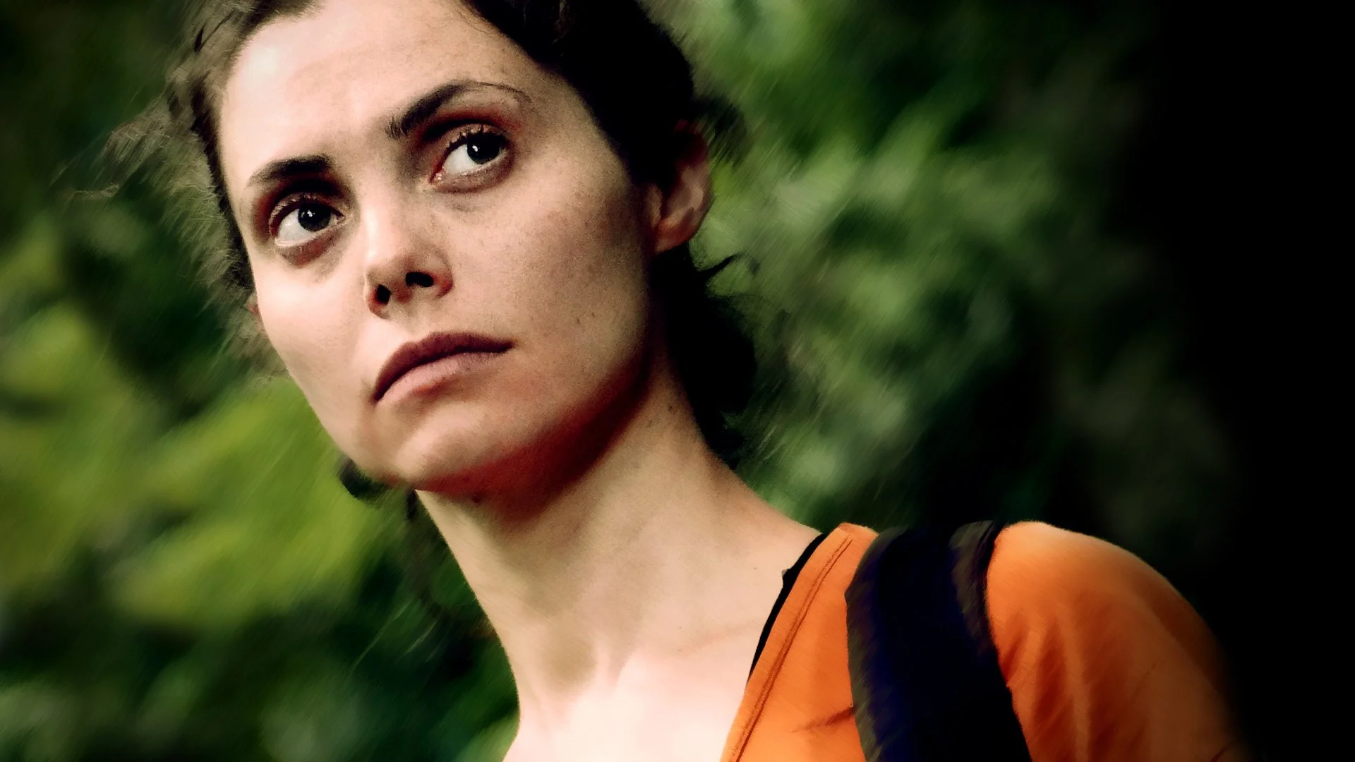 A young woman with dark hair pulled back, wearing an orange top and black backpack, standing outdoors with green foliage in the background, looking to the side with a serious expression.
