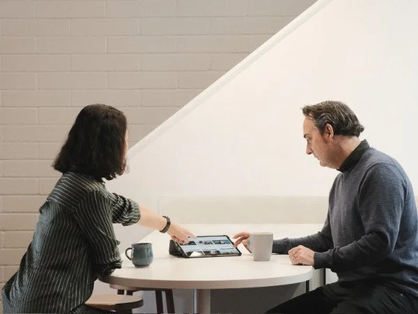 A woman and a man sitting at a round table, looking at a tablet, with coffee mugs in front of them, near a staircase and a brick wall.