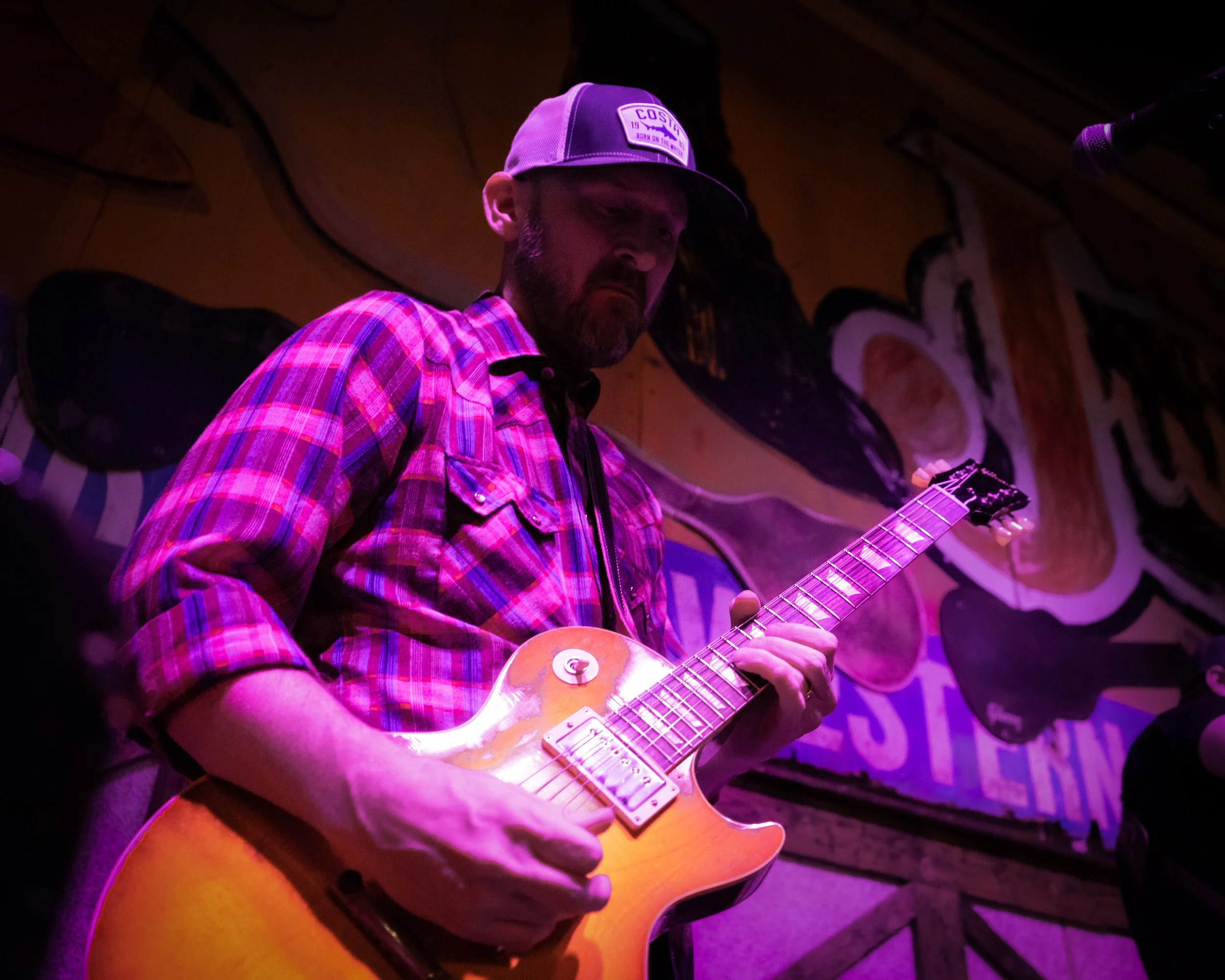 A man playing an electric guitar on stage with colorful lighting and a mural in the background.