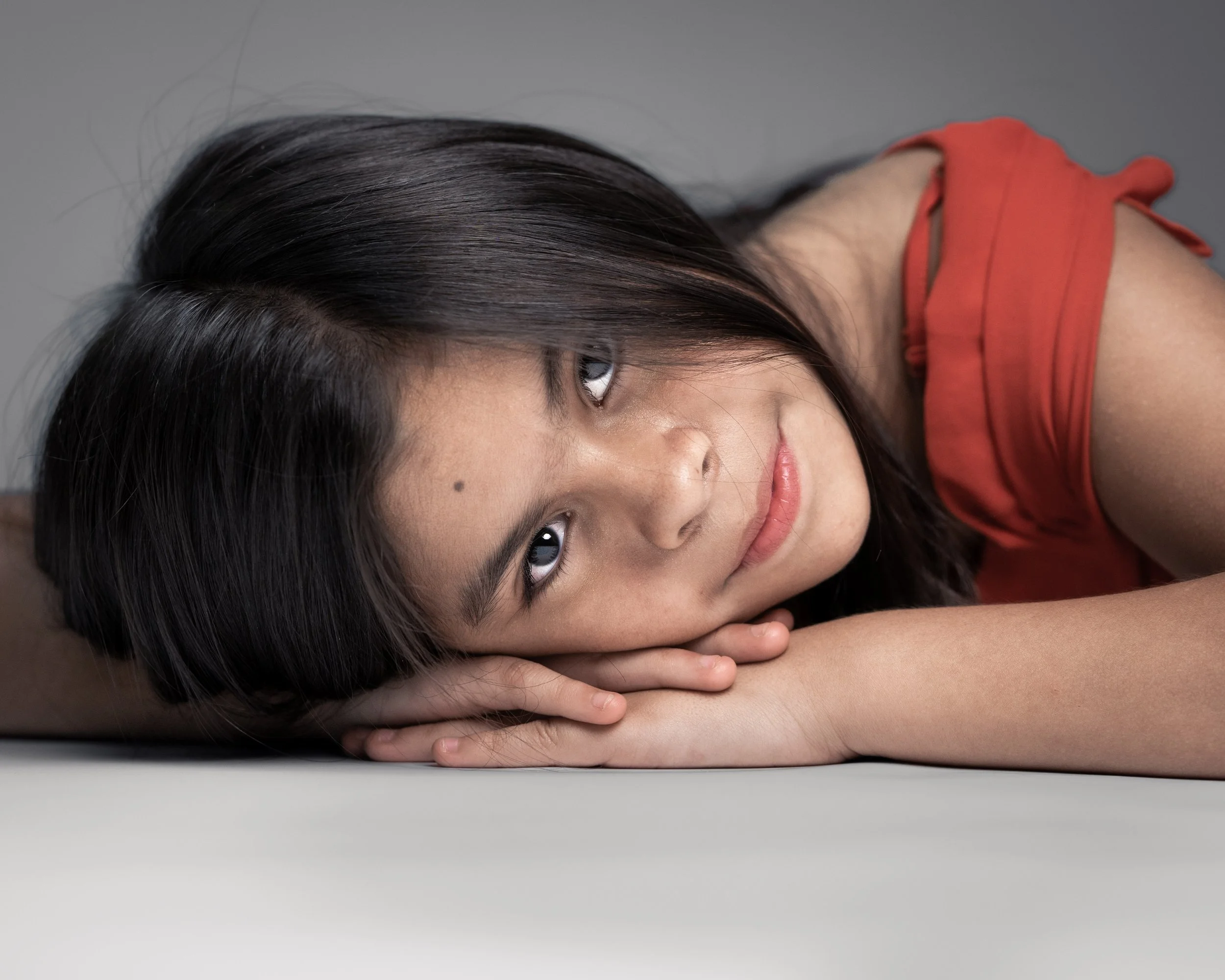 A young woman with dark hair lying on her side with her head resting on her arms, looking at the camera.