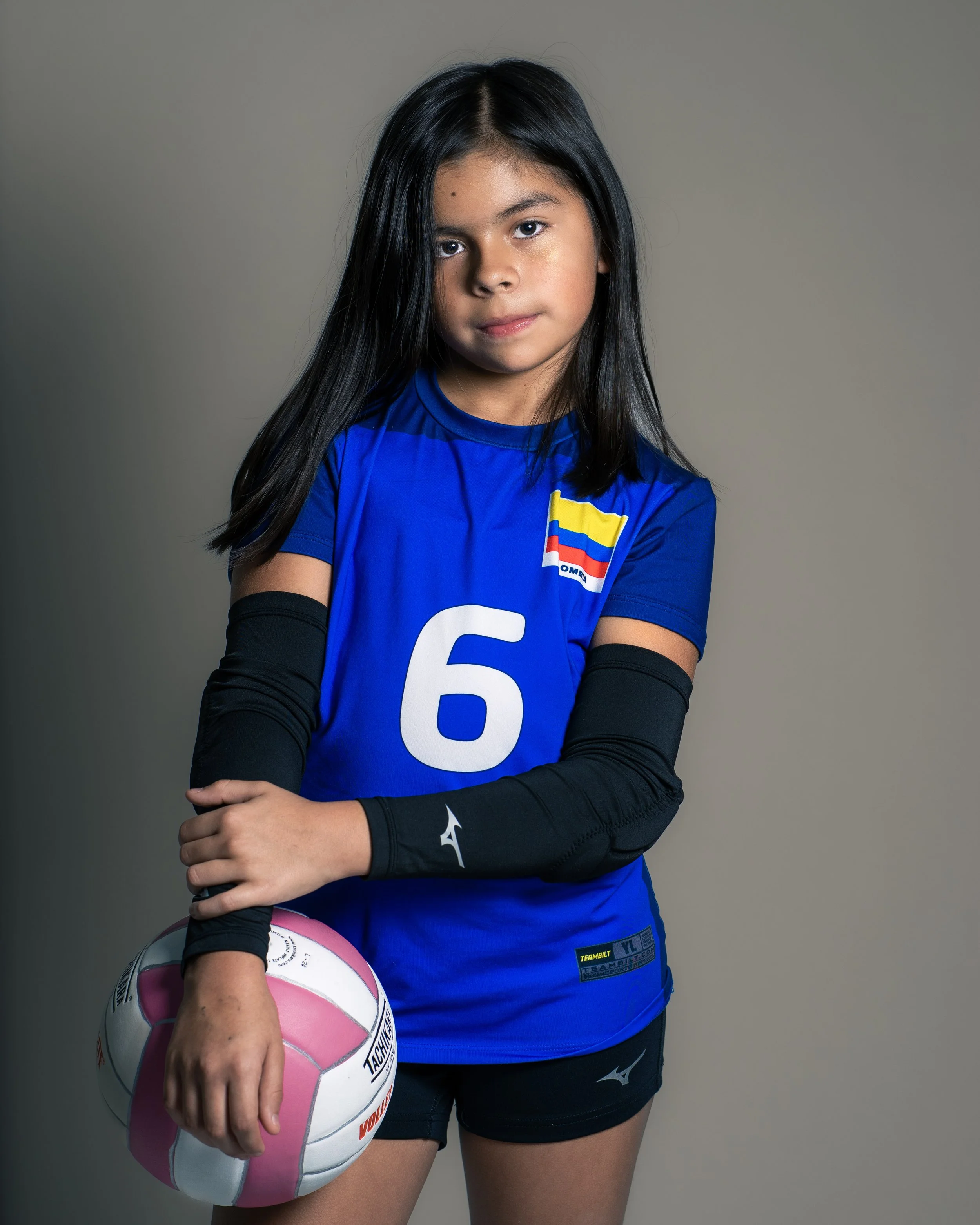 Young girl in volleyball uniform holding a pink and white volleyball against a gray background.