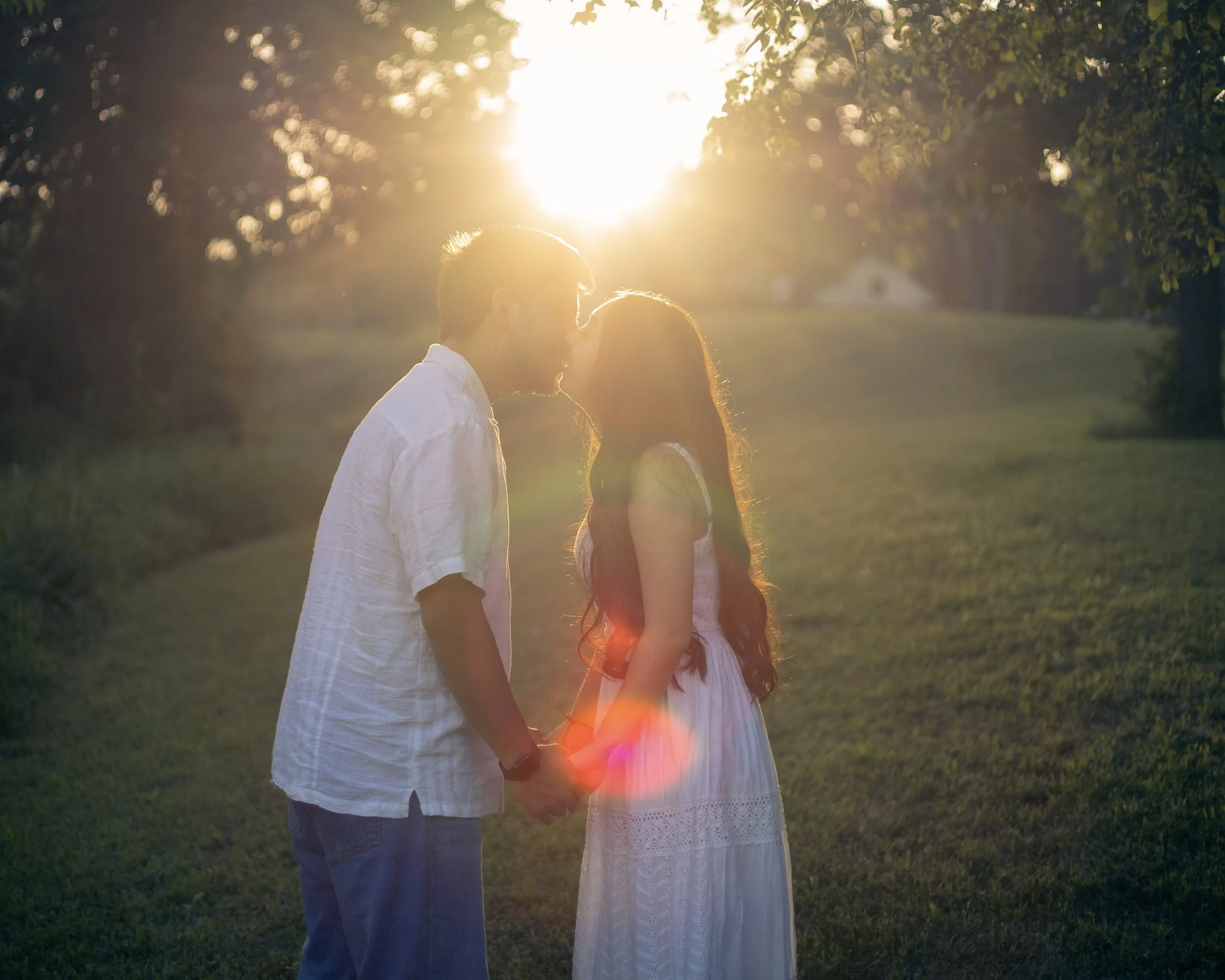 A couple holding hands, about to kiss, standing in a grassy field with trees in the background and the sun setting behind them.