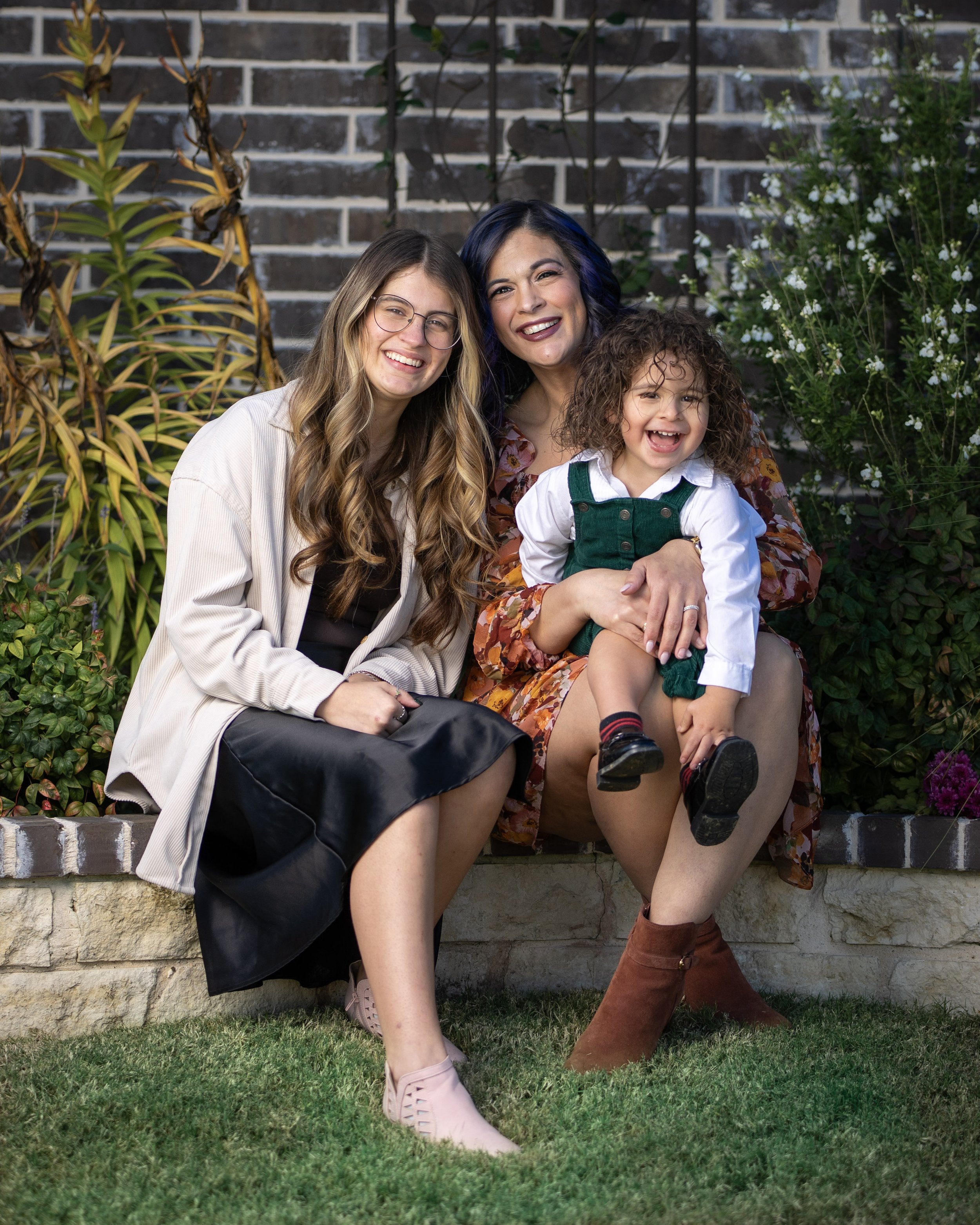 Three women and a boy sitting on a garden wall, smiling. They are surrounded by plants and a brick wall.