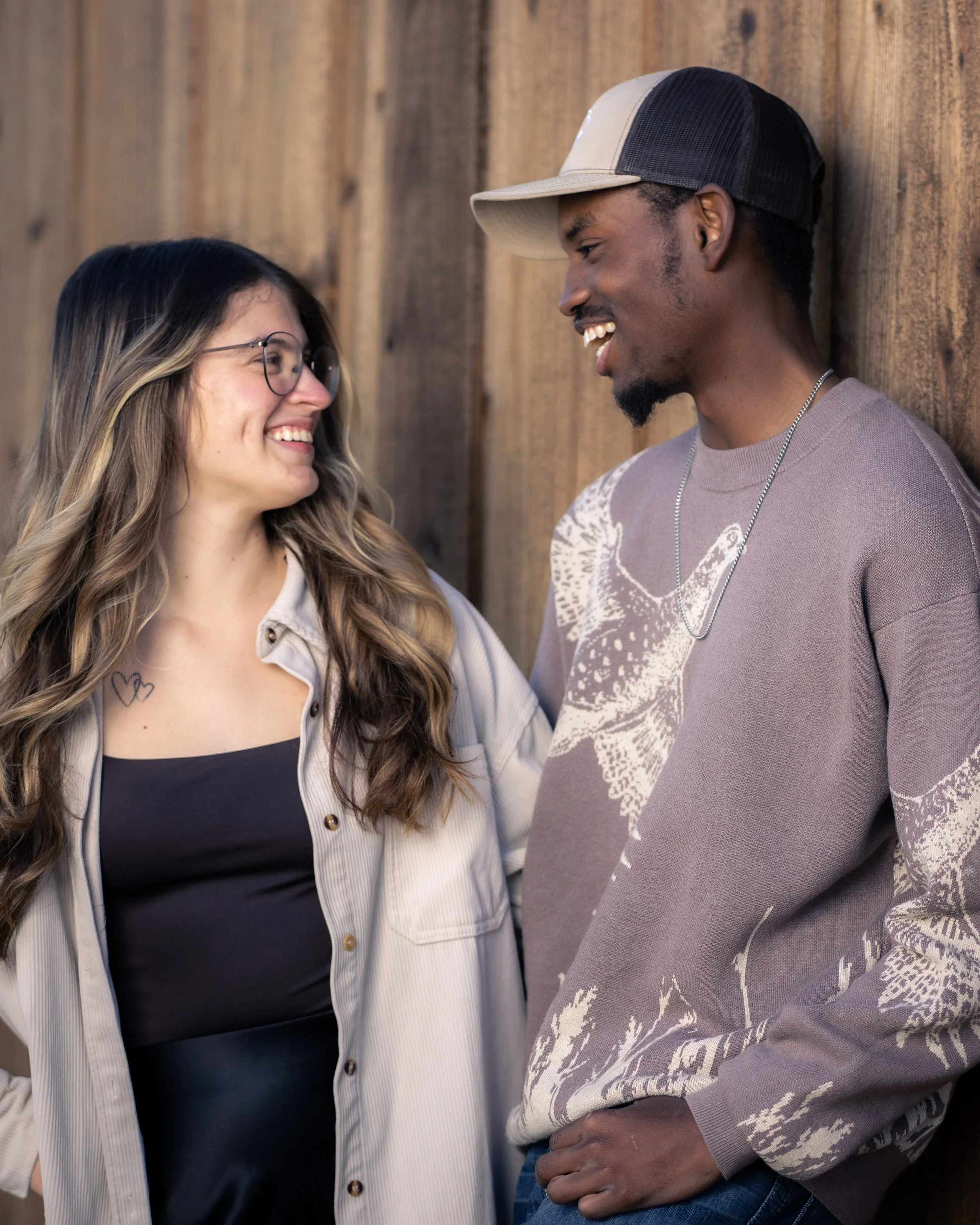A woman and a man smiling and looking at each other, standing against a wooden wall.
