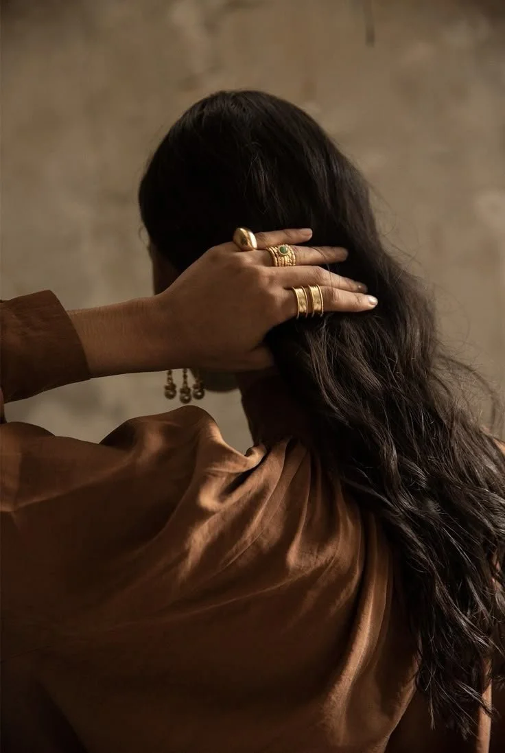 A woman with dark, wavy hair wearing multiple gold rings and earrings, dressed in brown clothing, gesturing with her hand on the back of her head, against a neutral background.