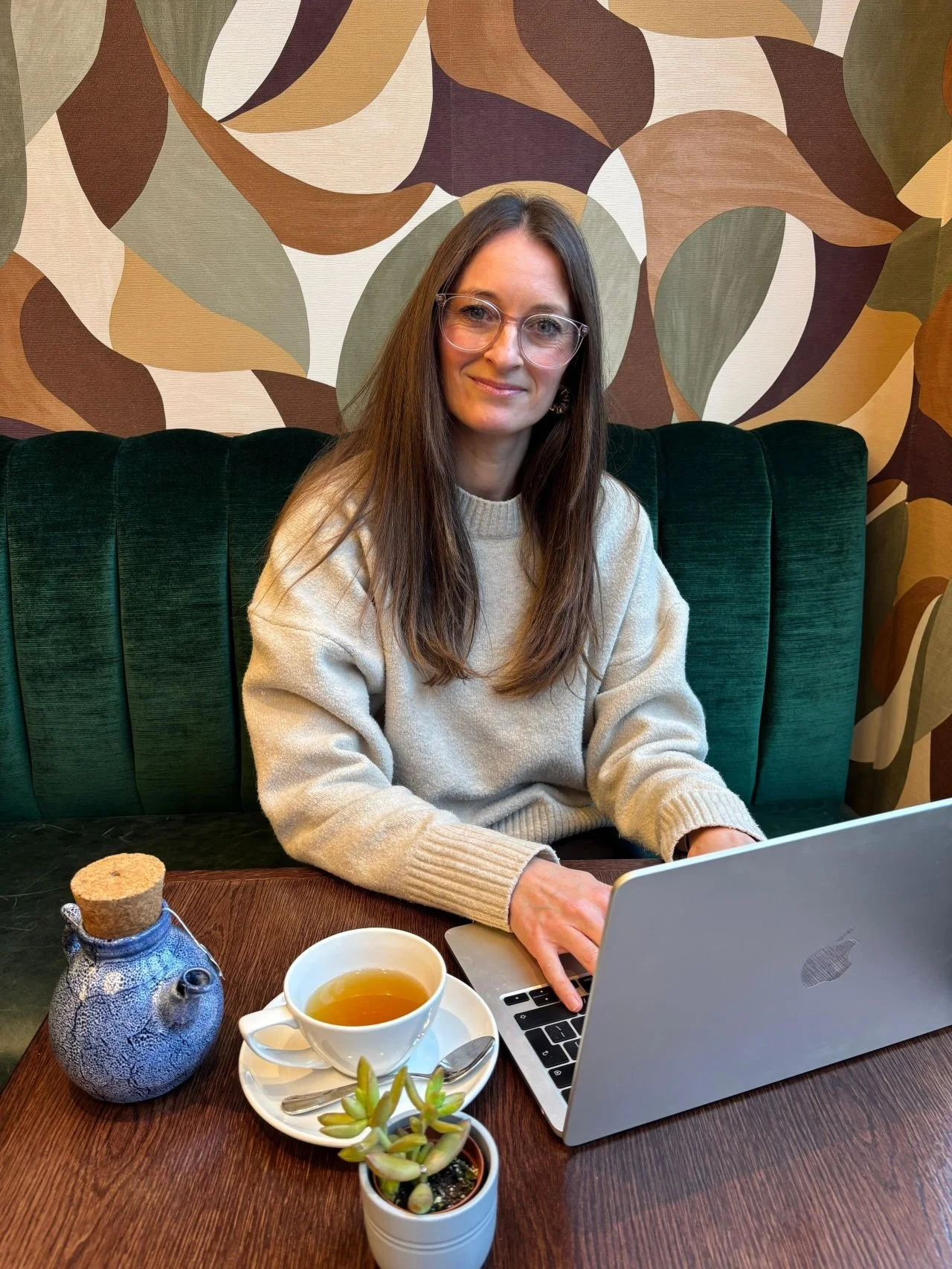 A woman with long brown hair and glasses sitting at a wooden table with a silver laptop, a cup of tea on a white saucer, a small potted succulent, and a blue ceramic teapot with a cork lid. She is wearing a beige sweater and is sitting in a green upholstered booth with a leaf-patterned wallpaper in the background.