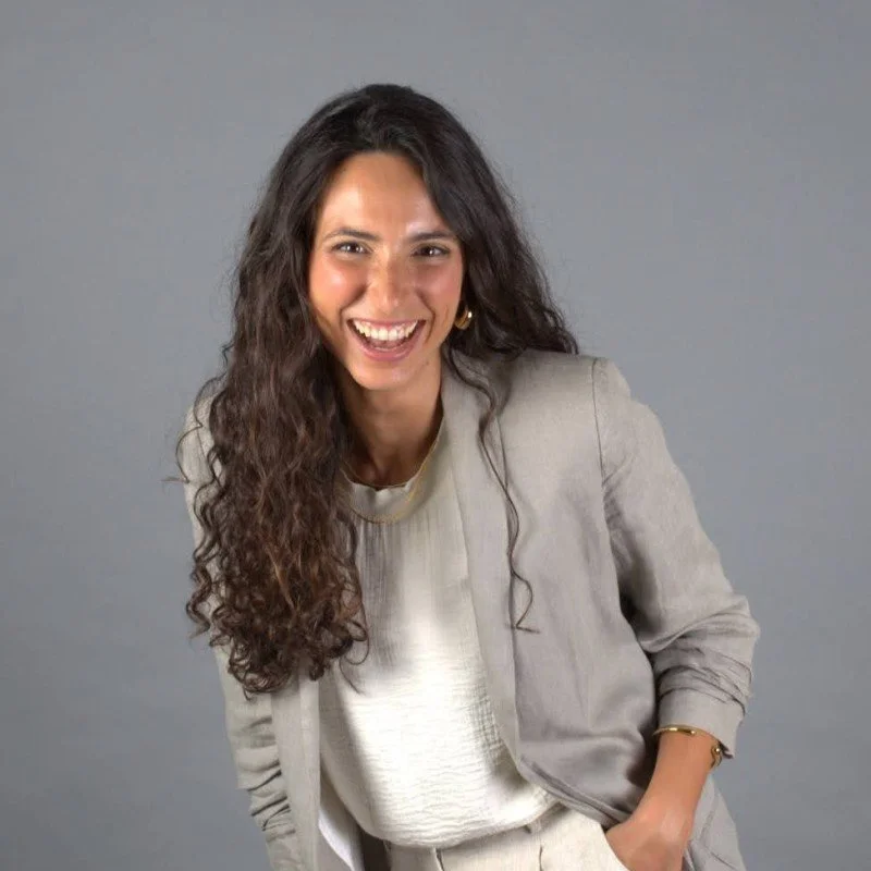 A woman with long, curly brown hair, smiling, wearing a light-colored blazer and blouse, standing against a plain grey background.