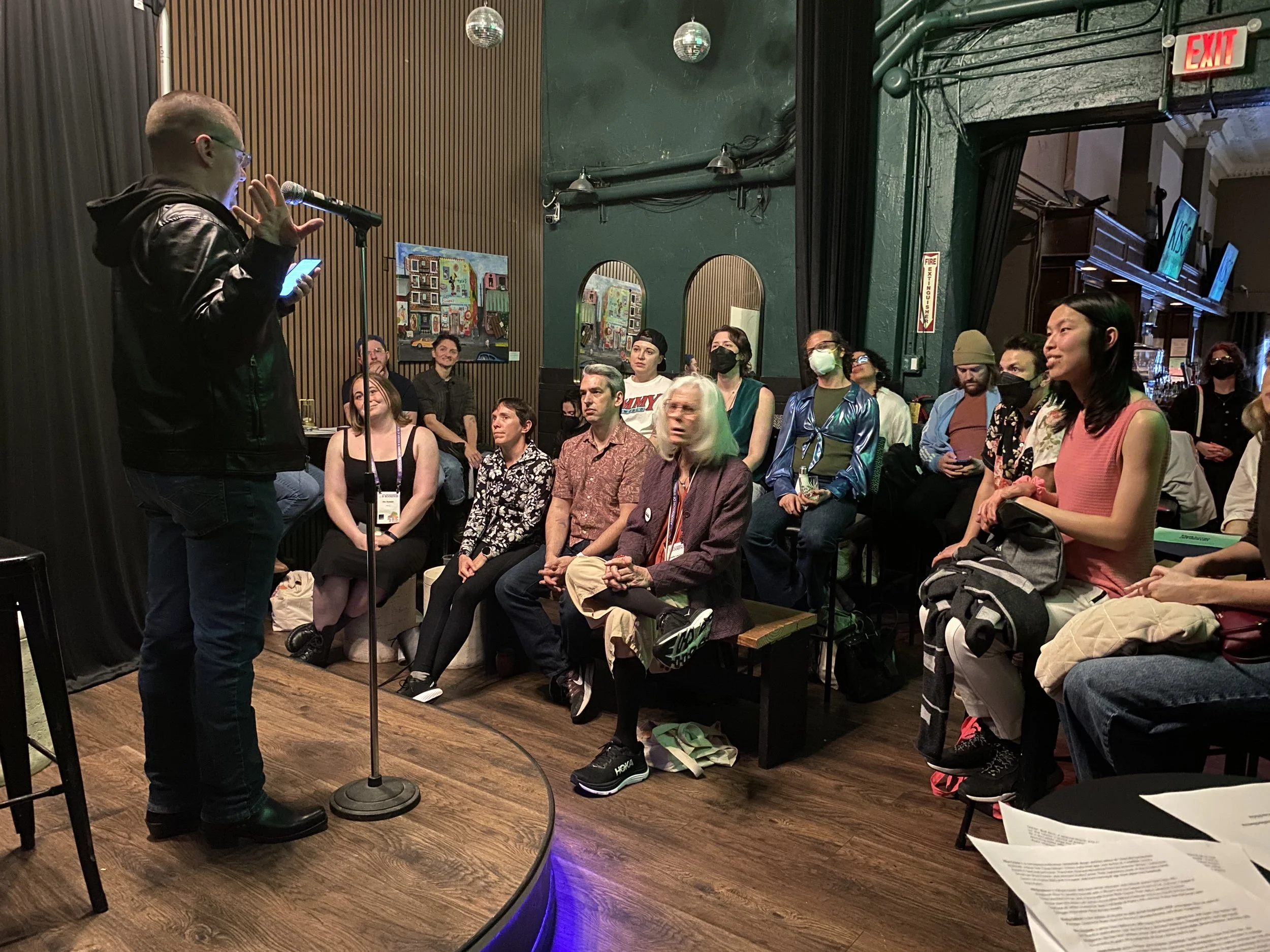 A man with glasses and a black leather jacket reading from a phone or card at a microphone while an audience listens in a dimly lit venue with dark green walls and artwork on the wall.