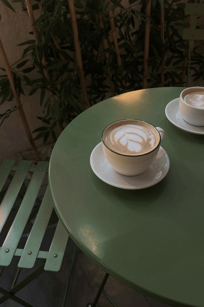 Two cups of latte on a green table with a plant in the background.
