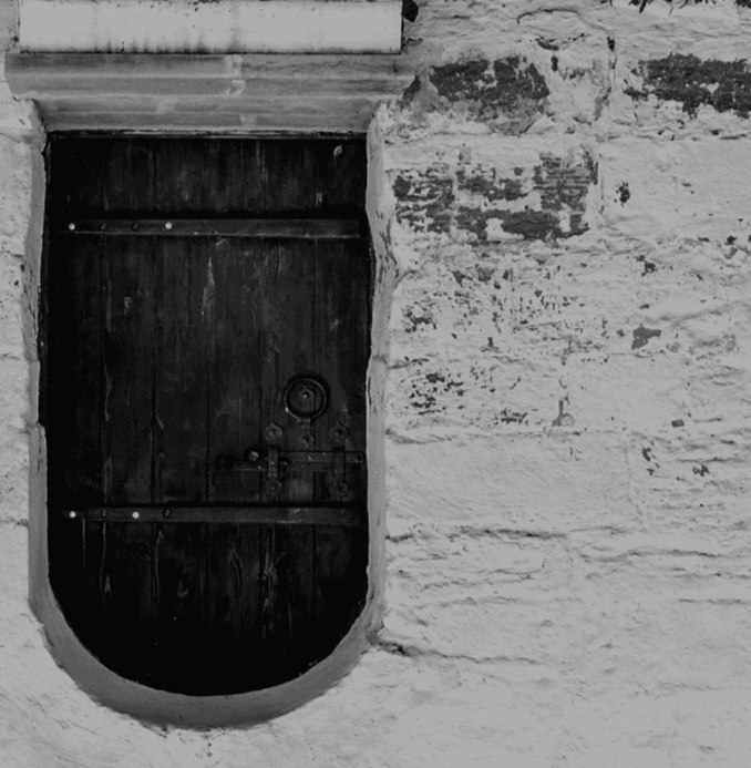 A small, dark wooden door with metal hinges and a small round doorknob, set into a white brick wall.