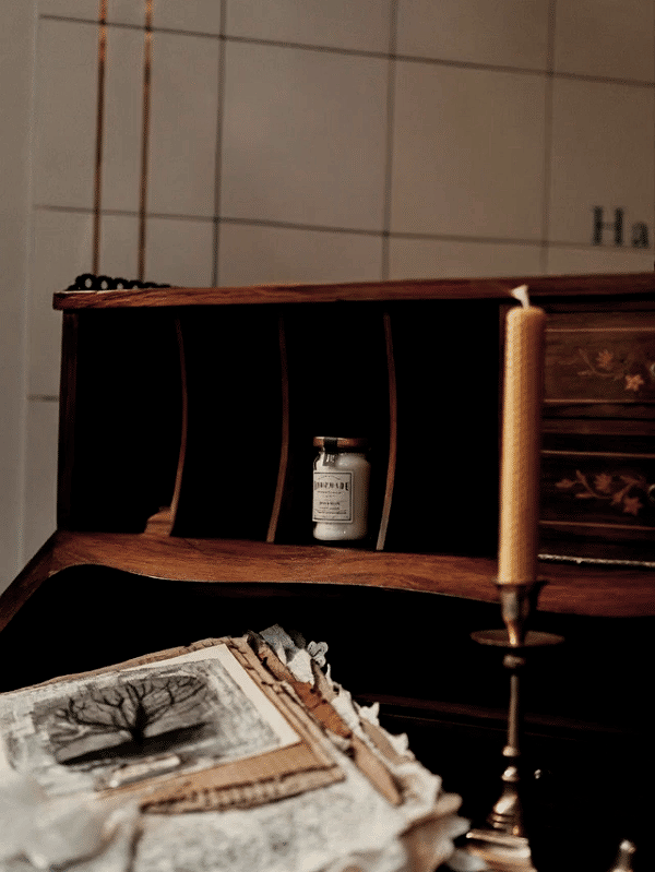 A wooden bookshelf with vertical compartments holding a small bottle, with a stack of papers or cloth with a picture of a tree in the foreground, and a candle holder with a candle on the right side.