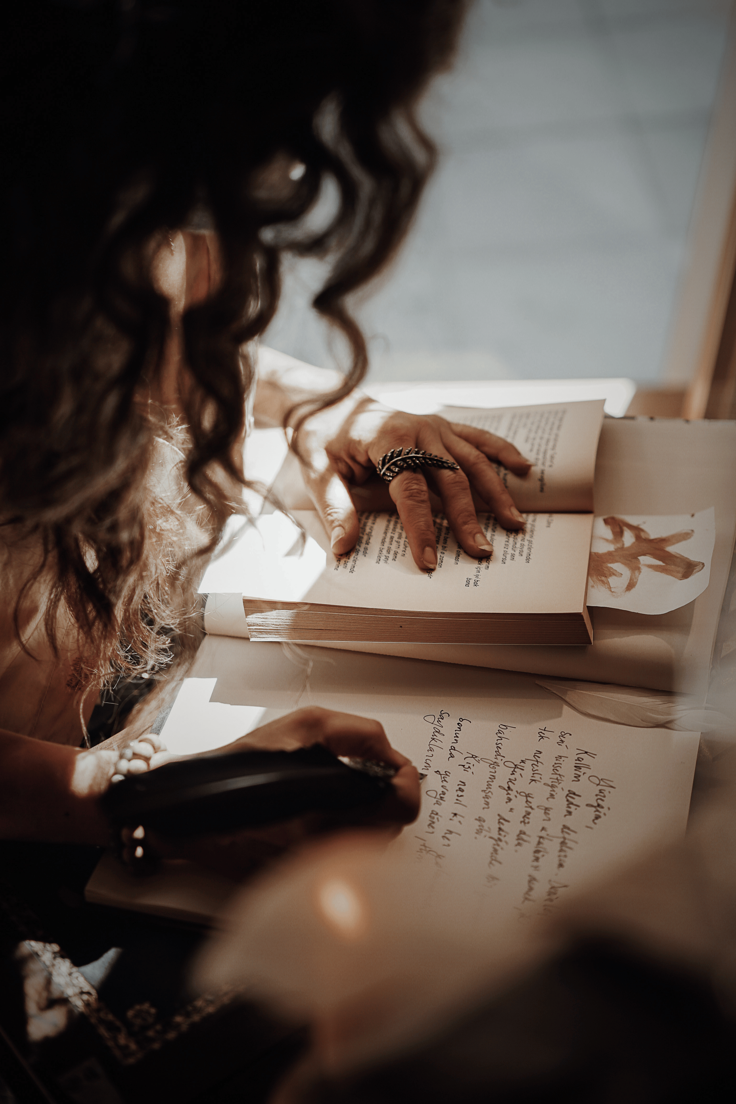 Close-up of a woman with curly dark hair reading a book, writing in a notebook with handwritten notes, in a well-lit room.