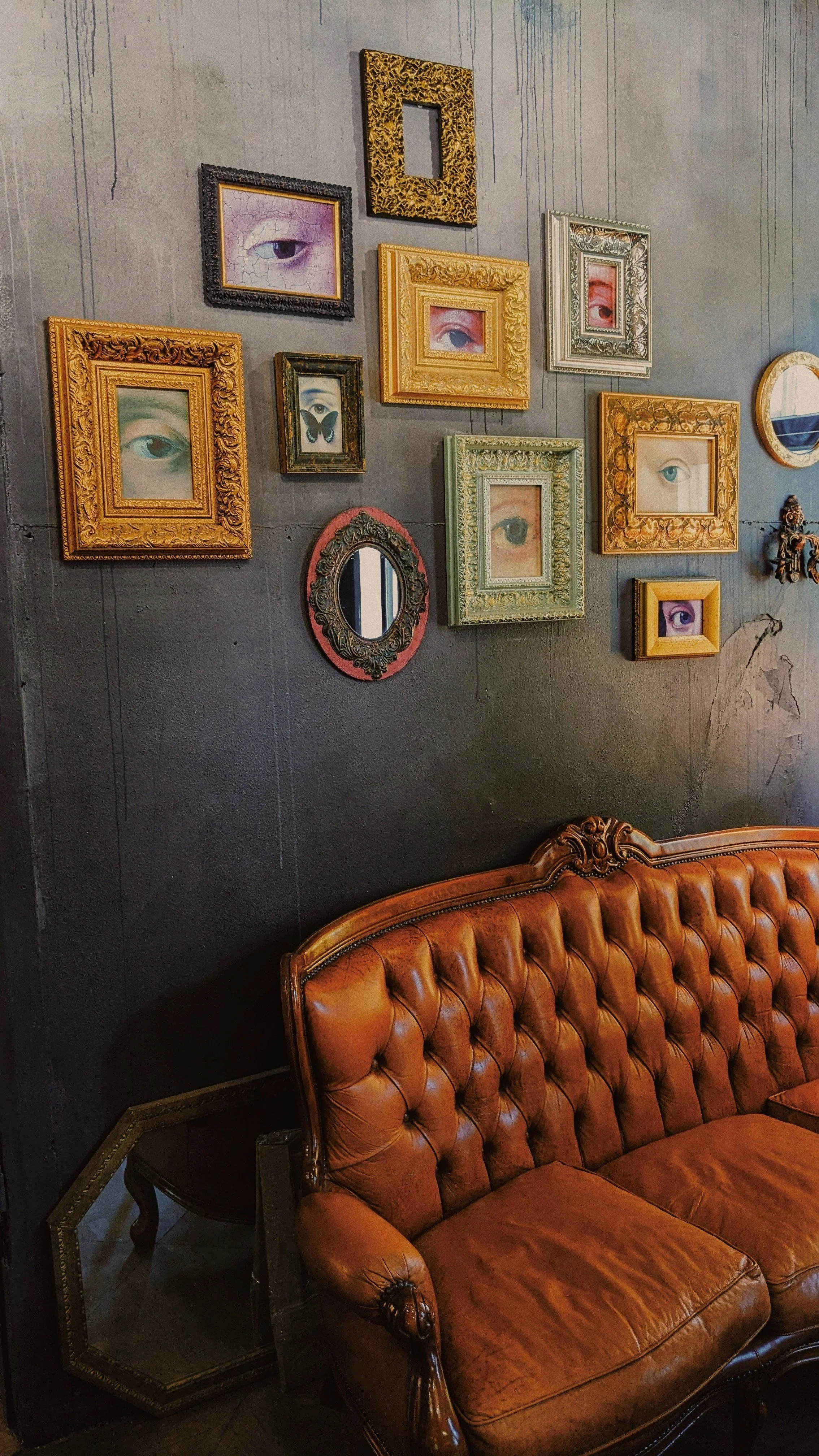 A dark gray wall decorated with multiple framed pictures of eyes and a butterfly, arranged in an eclectic gallery style. There is a vintage leather couch in front of the wall and a small mirror on a side table.