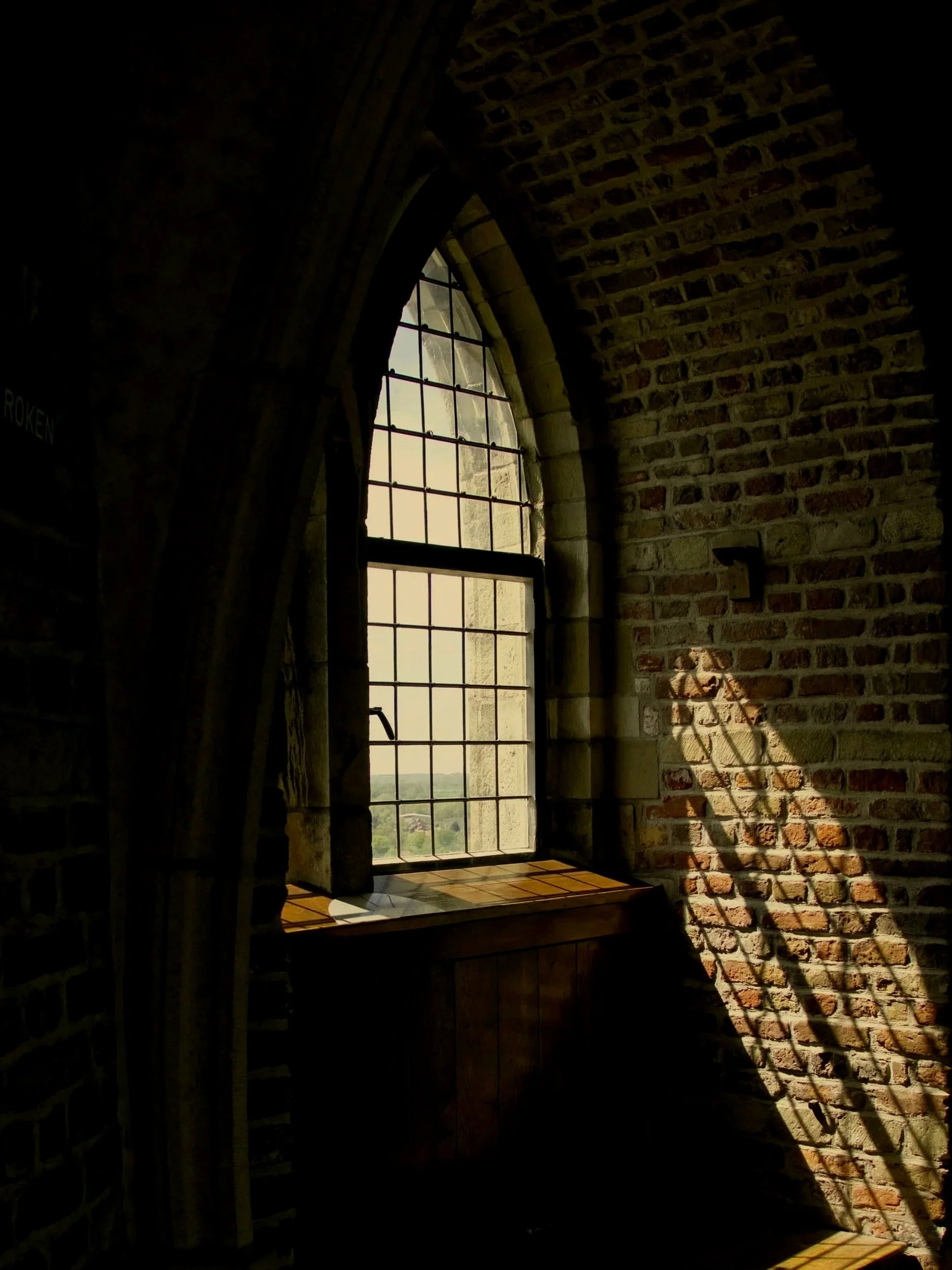 Sunlight streaming through a large arched window with metal grid panes, casting shadows on a brick wall inside a building.