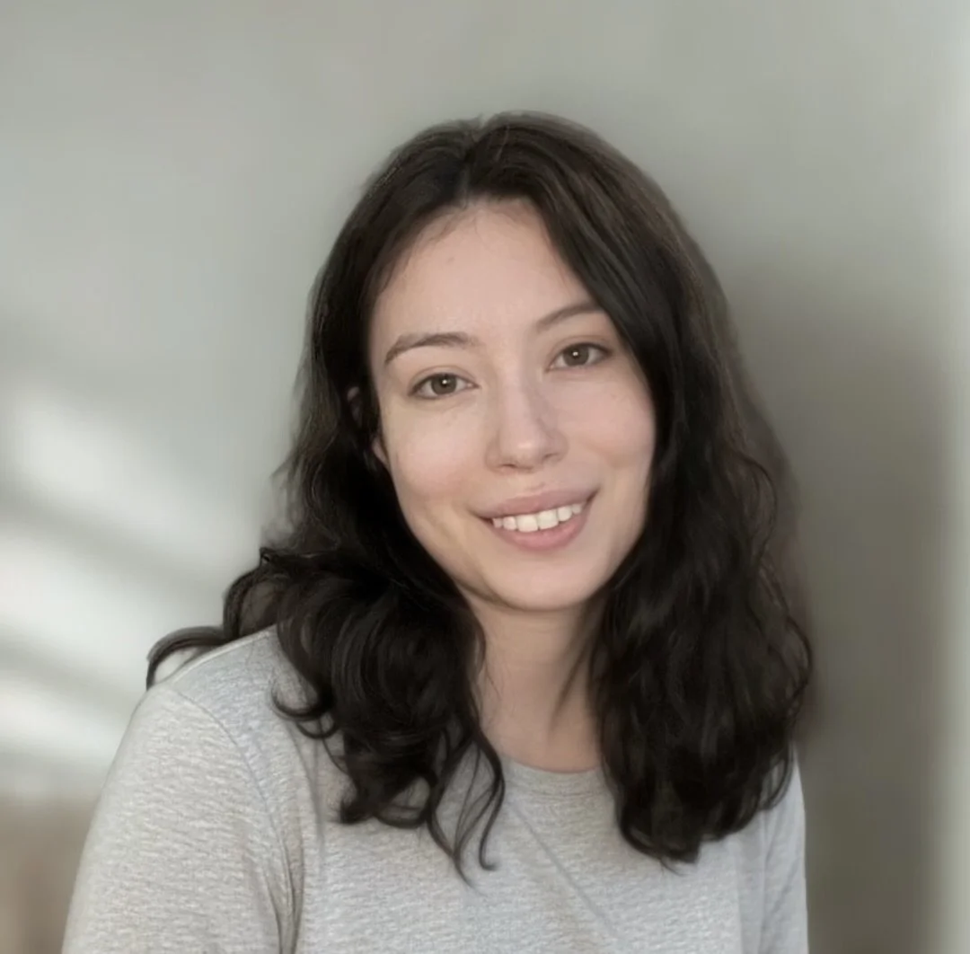 A young woman with long dark wavy hair smiling at the camera, wearing a light gray shirt, with a plain light-colored background.