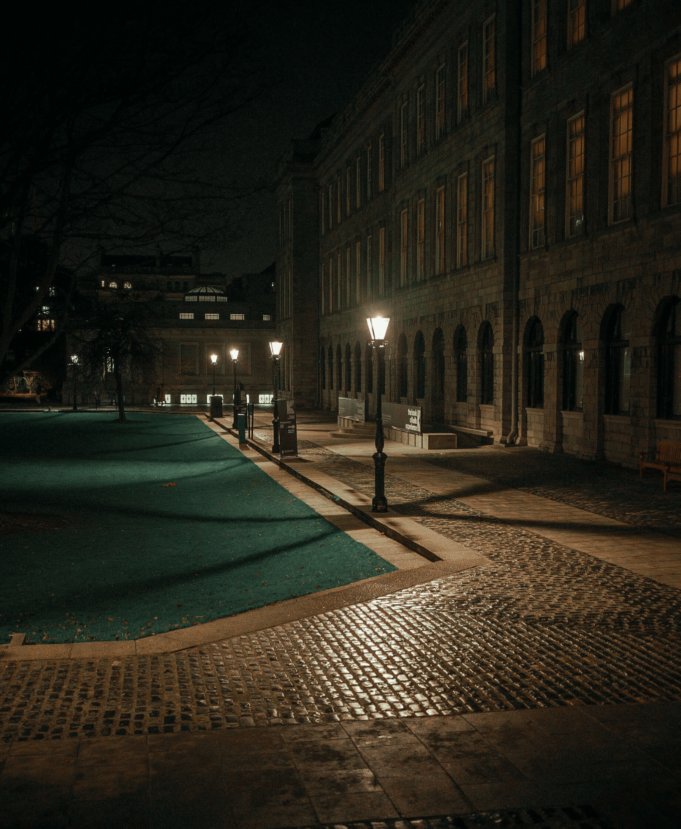 Nighttime scene of a sidewalk with vintage street lamps illuminating the pathway alongside a large stone building with multiple windows.