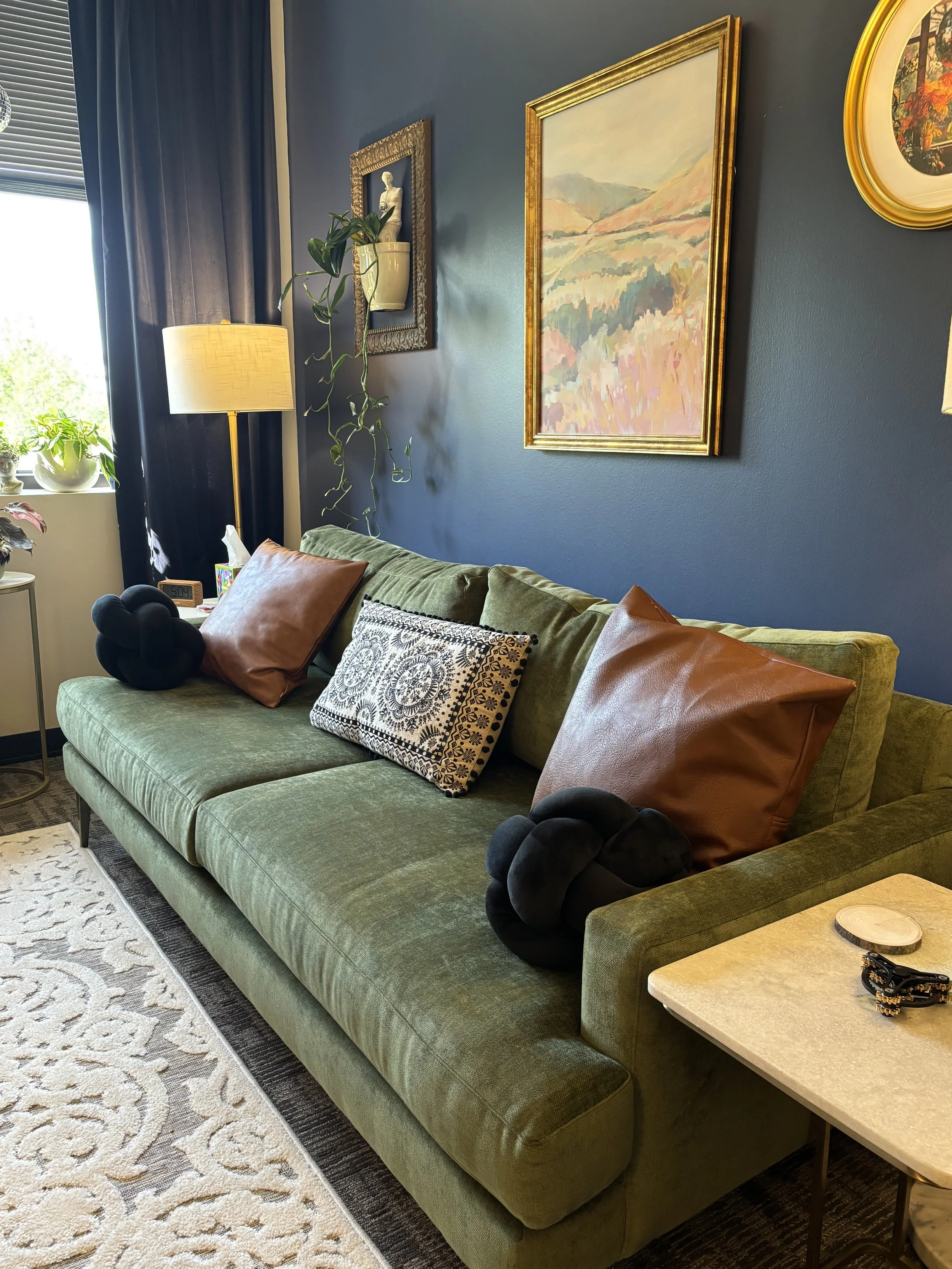 Living room with a green velvet sofa adorned with brown and patterned pillows, a side table with decorations, and art on a blue wall.