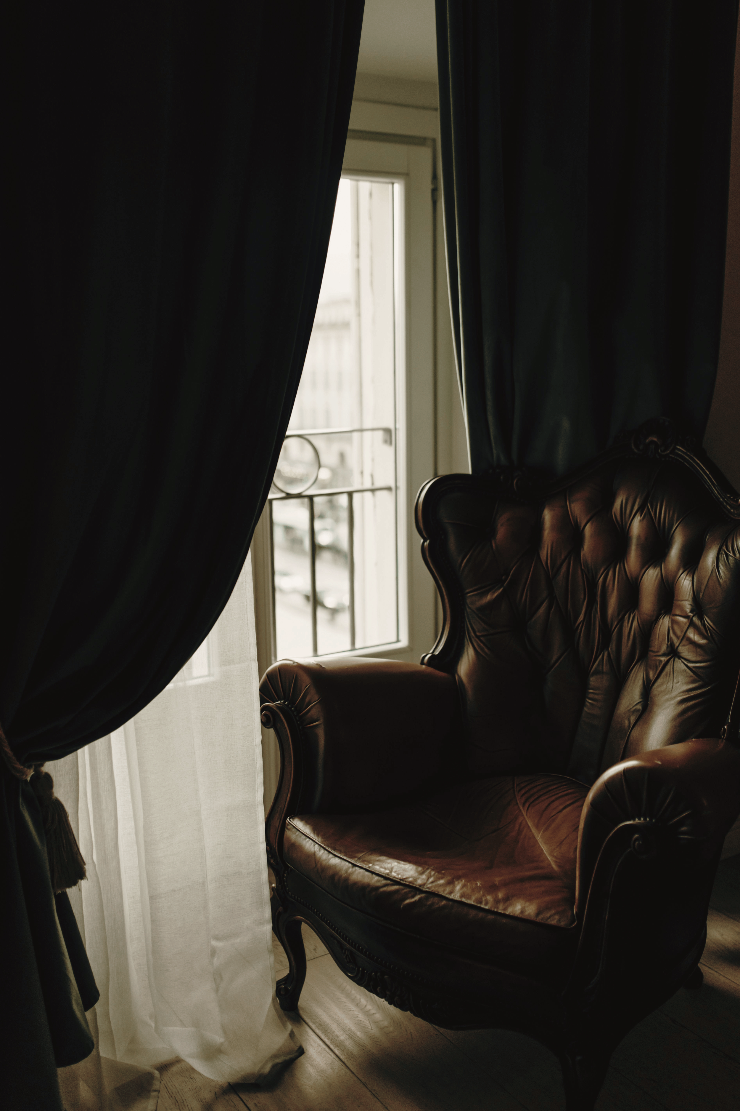 A vintage leather armchair situated next to a window with sheer white and dark curtains, allowing natural light into the room.