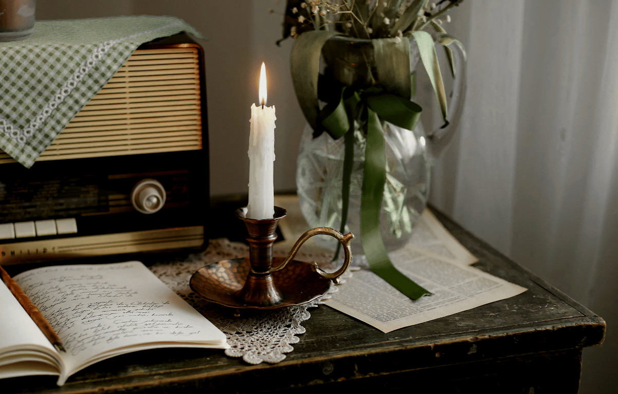 A lit candle in a vintage brass holder on an antique wooden table, with a flowered book, open notebook, and glass vase with flowers in the background.