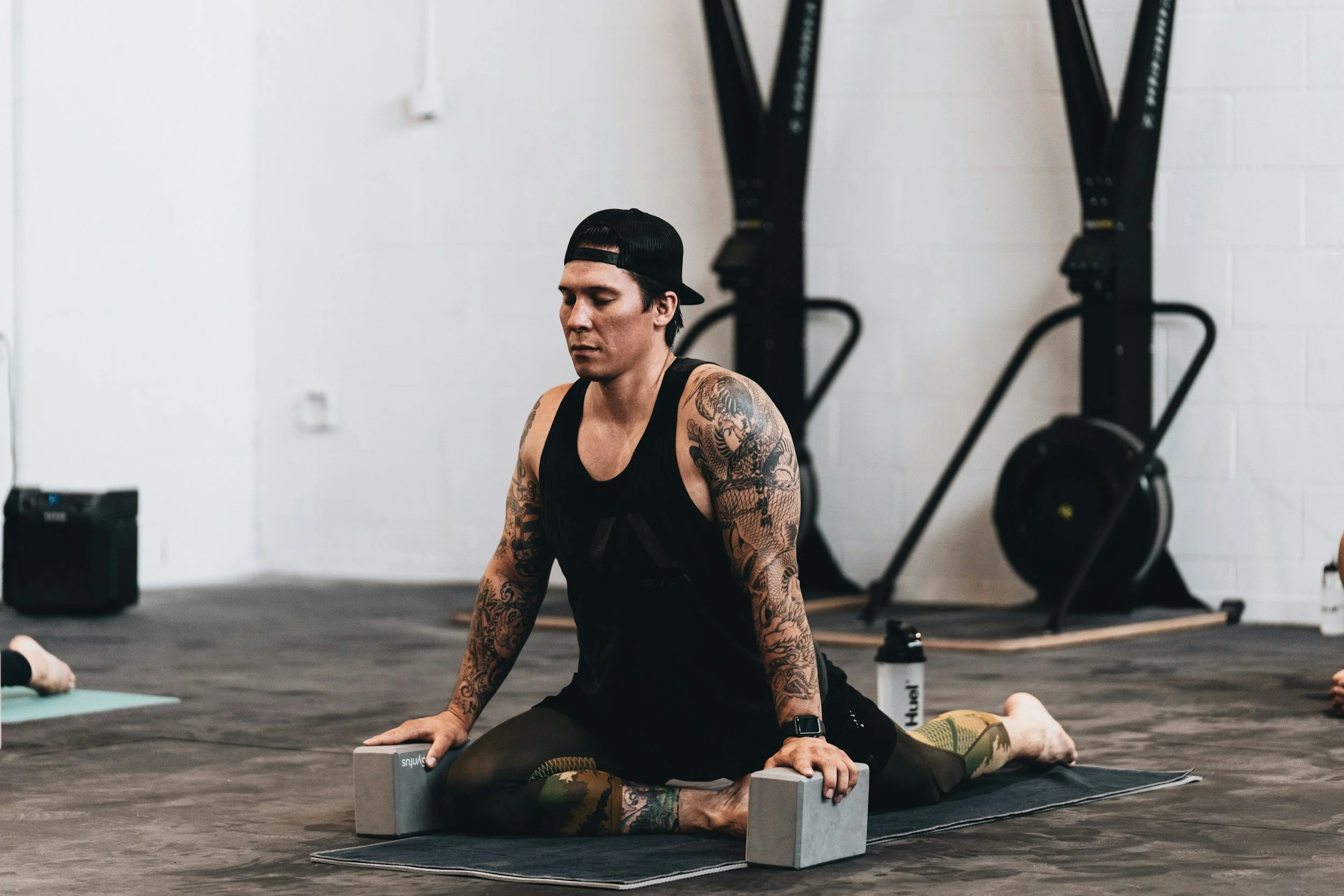 Person with tattoos practicing yoga or stretching in a gym, using yoga blocks and mats, with workout equipment in the background.