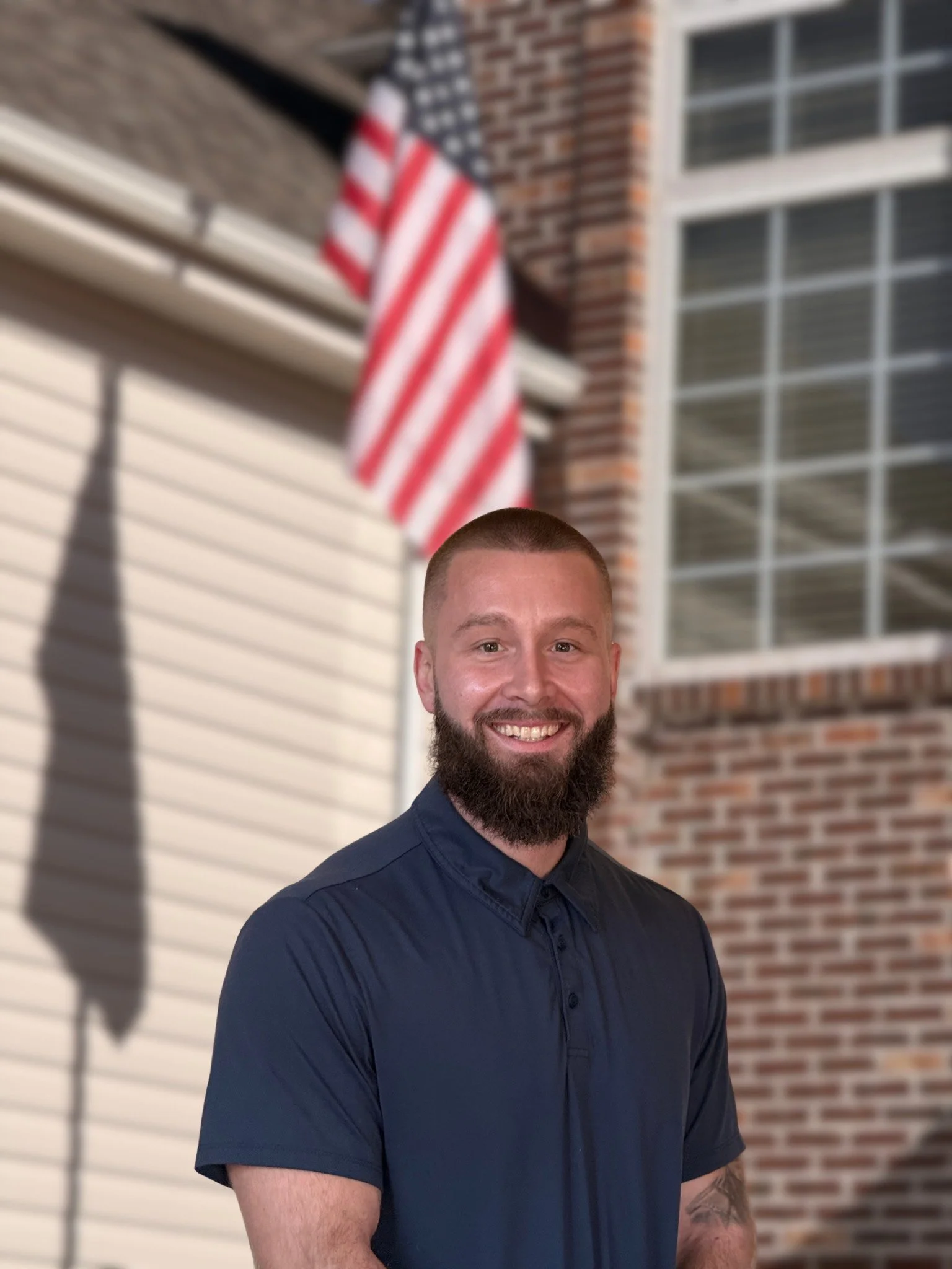 A smiling man with a beard and short hair standing outside near a brick house with a window. An American flag is hanging behind him. The house has beige siding and a shutter.