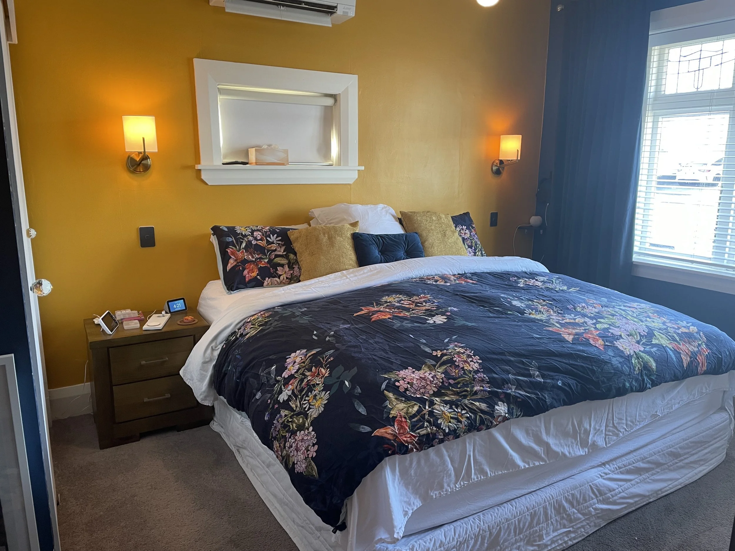 A bedroom with a yellow accent wall, a bed with floral bedding and multiple pillows, nightstands with reading lamps, a window with blinds and curtains, and a wall-mounted light.