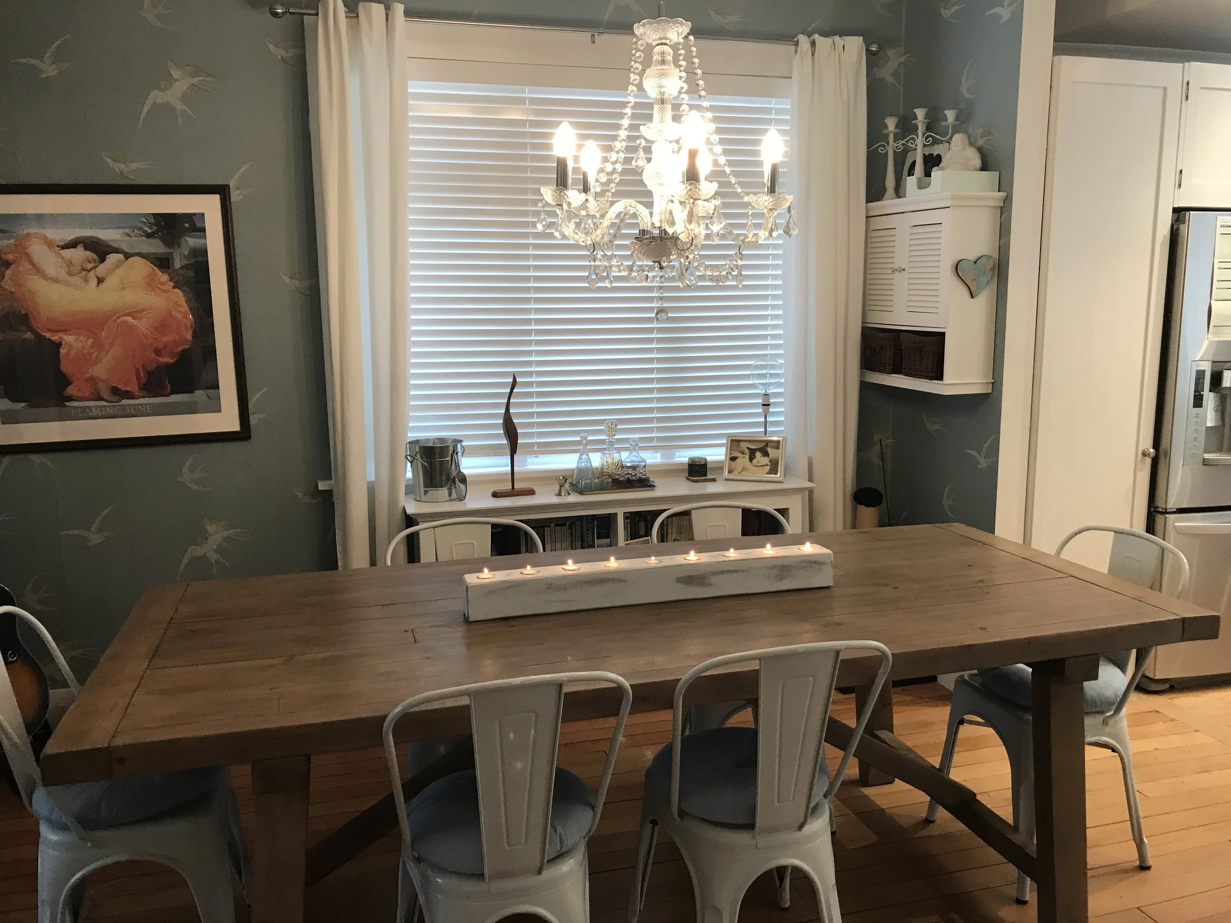 A dining room with a wooden table and six white metal chairs, a chandelier hanging above, a window with white blinds and curtains behind the table, decorated with candles, candles holder, picture frames, and decorative objects.